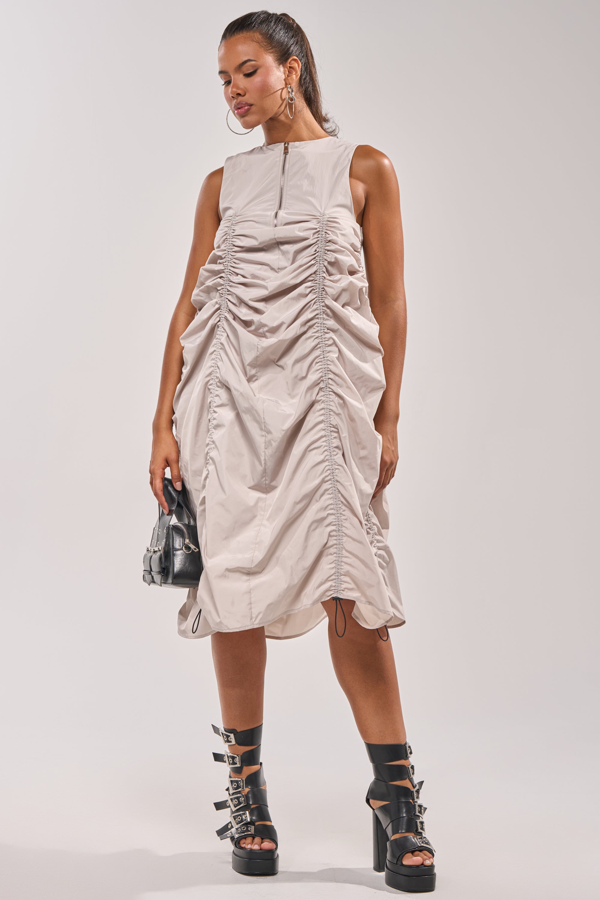A woman in the SORRY NOT SORRY NYLON CINCH MINI DRESS, styled with black strappy heeled boots, large hoop earrings, and a small black handbag, poses against a plain background. 10
