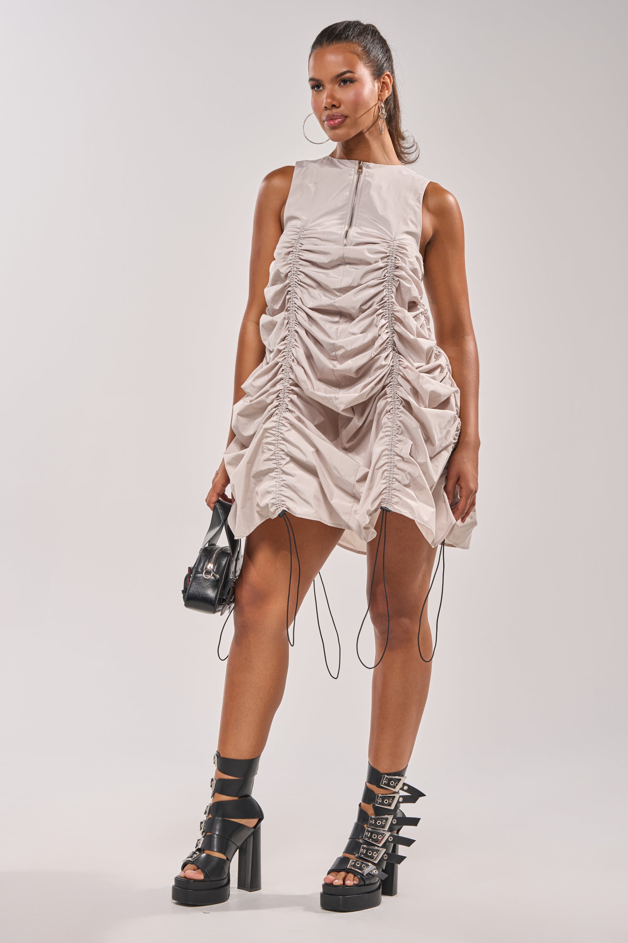 A woman stands confidently in a street-style look, wearing the SORRY NOT SORRY NYLON CINCH MINI DRESS with drawstring details, black platform sandals, a small black handbag, hoop earrings, and her hair styled back. 11