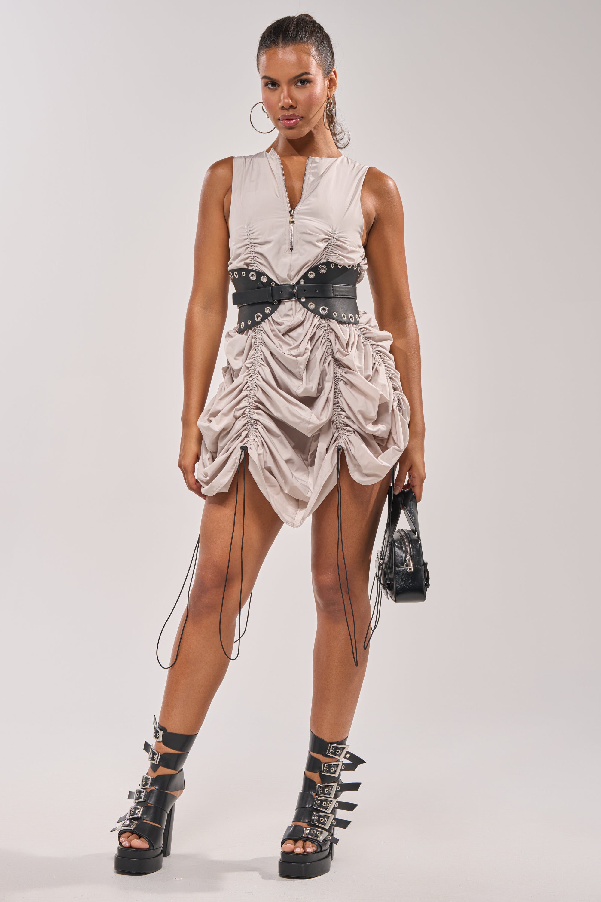 A woman stands confidently against a plain background in the SORRY NOT SORRY NYLON CINCH MINI DRESS, styled street-fashion with a black studded belt, small black bag, and black strappy platform boots. 12