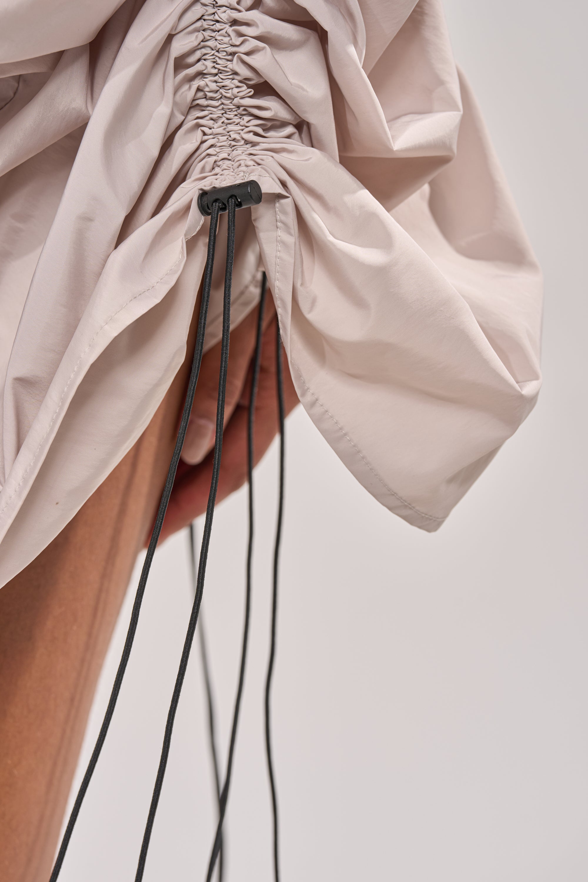 Close-up of the SORRY NOT SORRY NYLON CINCH MINI DRESS in beige. The lightweight, slightly shiny fabric is gathered with black drawstrings and a plastic toggle, capturing modern street-style against a neutral background. 16