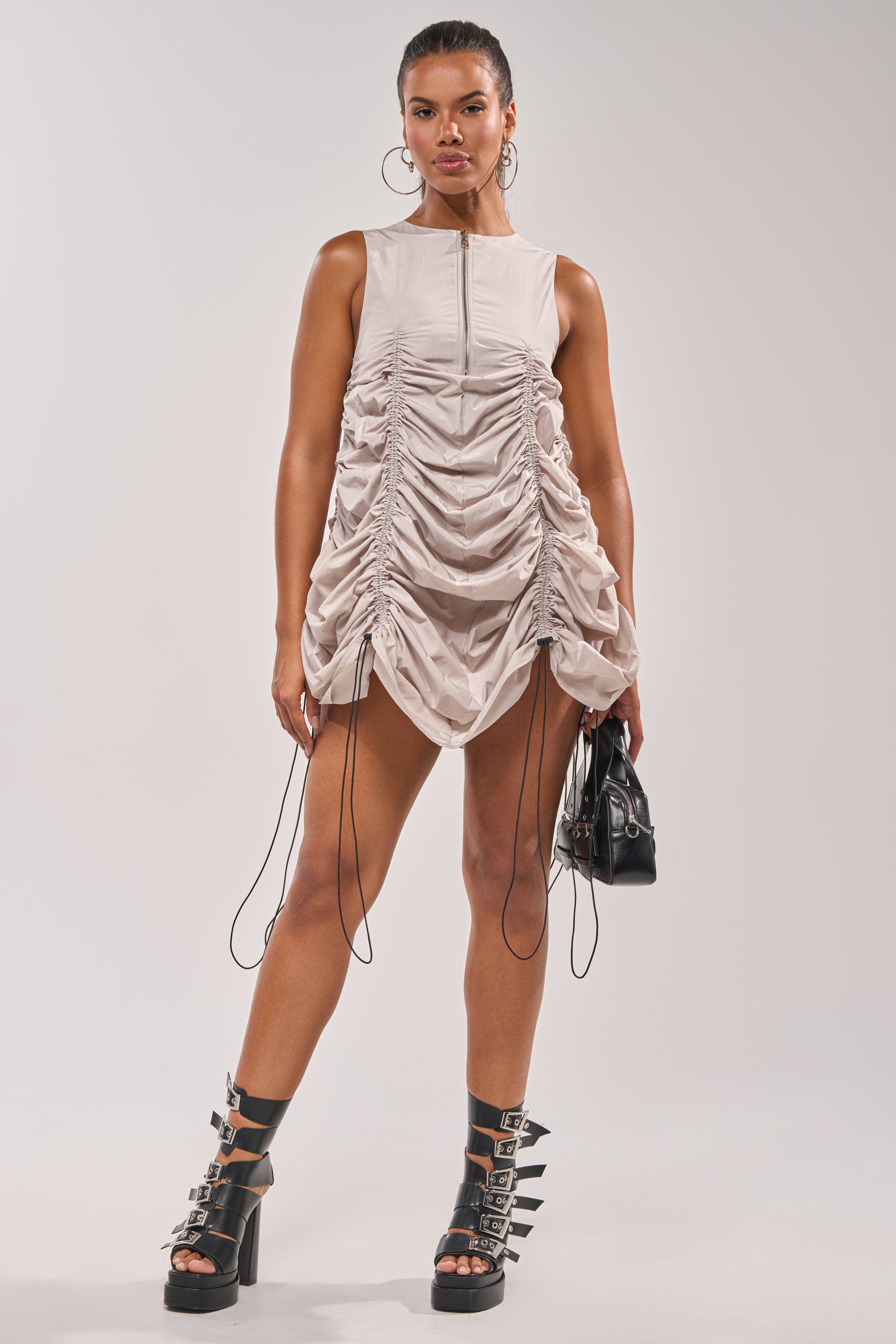 A woman stands confidently in the SORRY NOT SORRY NYLON CINCH MINI DRESS with drawstrings, paired with black buckle platform boots, large hoop earrings, and a small black handbag against a plain light background. 7
