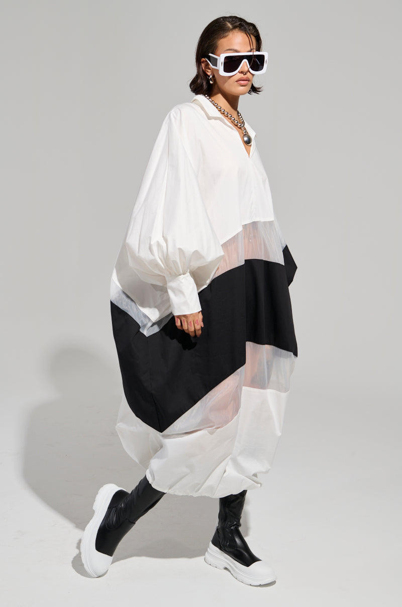 A person dressed in the DAY AND NIGHT COLORBLOCKED MIDI DRESS, featuring sheer mesh sections with black and white colorblocking, is wearing oversized sunglasses and black knee-high boots. They accessorize with a chunky necklace while striding confidently against a plain background.