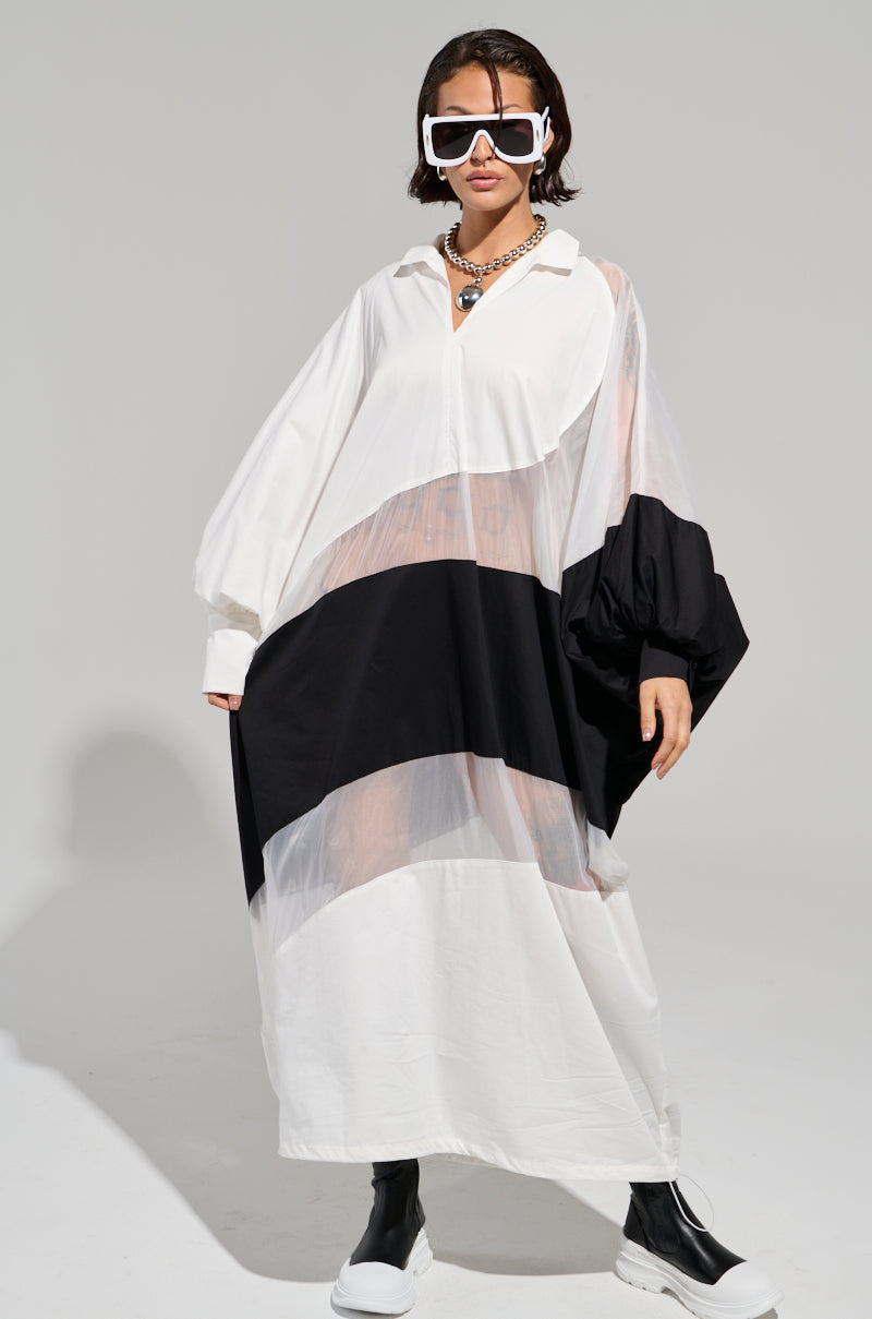 Wearing oversized white sunglasses and a bold statement necklace, this confident individual captivates in the DAY AND NIGHT COLORBLOCKED MIDI DRESS, marked by its sheer mesh sections. They finish the ensemble with chunky black and white boots against a simple, neutral gray background.