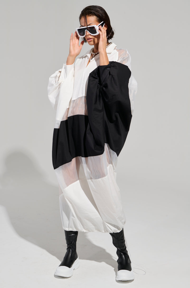 Wearing the chic DAY AND NIGHT COLORBLOCKED MIDI DRESS, featuring a stylish and oversized black and white design with an adjustable drawstring hem, a person strikes a confident pose. They adjust their white-framed sunglasses while their black boots complete the look against a plain background that highlights the dress's elegant details.