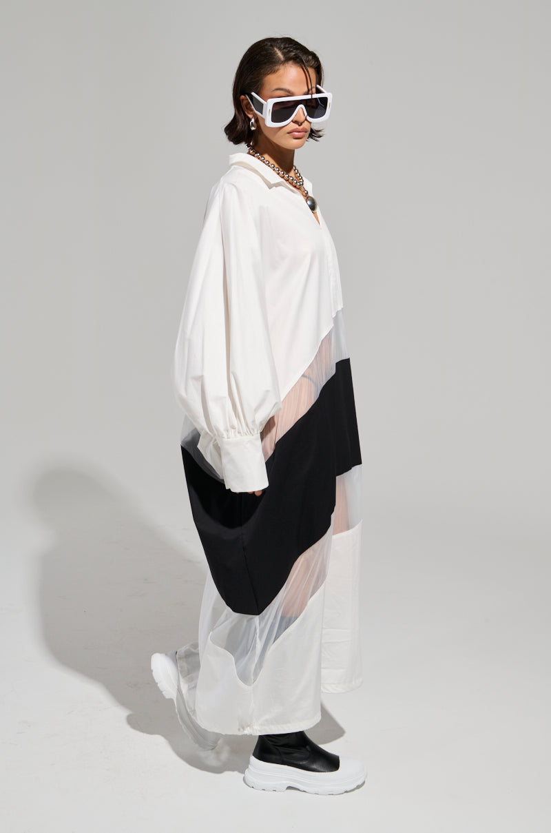 A model poses against a plain background, wearing the DAY AND NIGHT COLORBLOCKED MIDI DRESS, which boasts a flowing design with layers incorporating sheer mesh elements and adjustable drawstring hems. The look is completed with oversized sunglasses and white platform shoes.