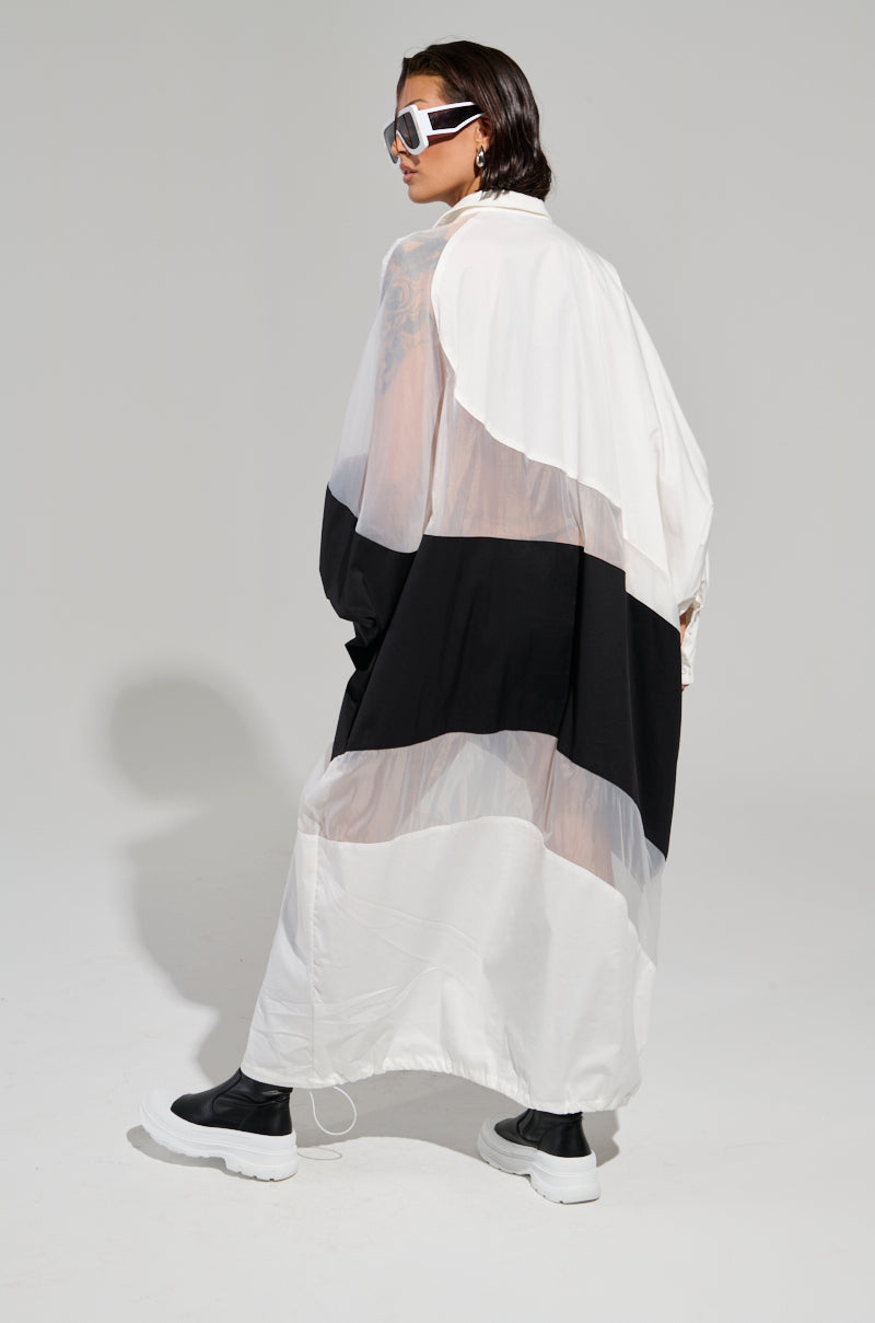 A person wearing a chic DAY AND NIGHT COLORBLOCKED MIDI DRESS, designed with an adjustable drawstring hem, stands against a plain background. They have short hair, sunglasses, and black ankle boots, looking sideways with a confident posture.
