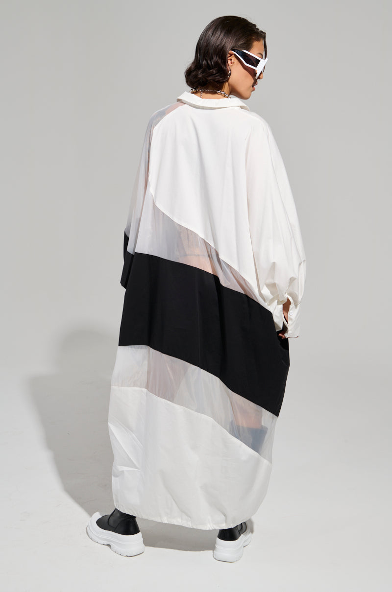A person wearing the DAY AND NIGHT COLORBLOCKED MIDI DRESS, featuring an oversized design with long sleeves and black and white geometric patterns, stands in a neutral studio setting. The dress includes an adjustable drawstring hem. They accessorize with sunglasses and black shoes while facing away from the camera.