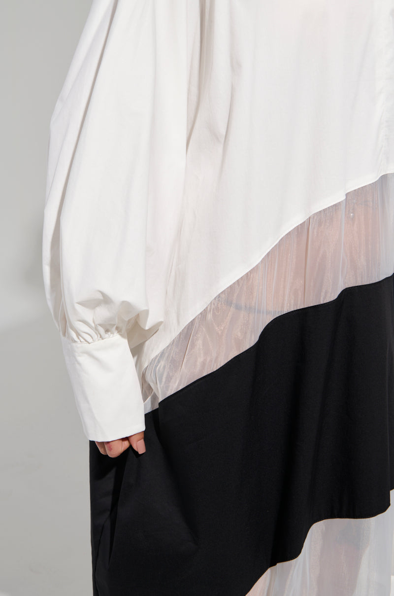 A person wears the DAY AND NIGHT COLORBLOCKED MIDI DRESS, an oversized white shirt with billowy sleeves and an asymmetrical design. It features sheer mesh accents and a black section at the bottom, offering a hint of elegance reminiscent of a layered garment.
