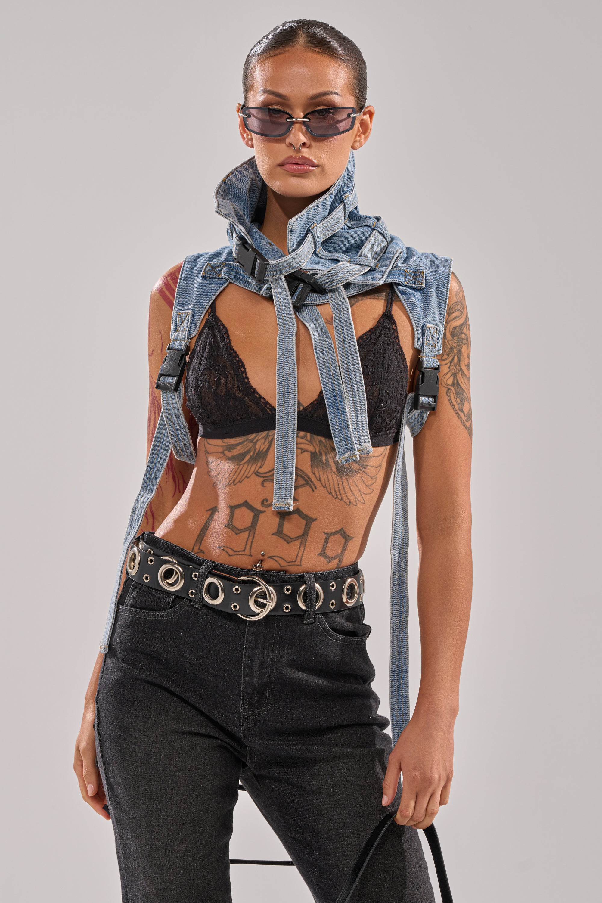A woman with tattoos, slicked-back hair, sunglasses, and a confident look wears a black lace bralette, black jeans with a grommet belt, and the NO SHOW JACKET COLLAR draped over her shoulders.