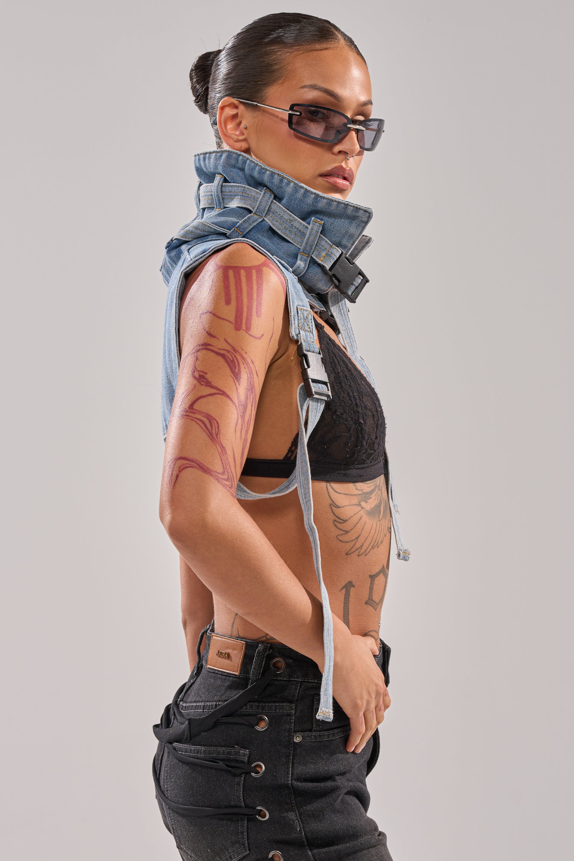 A woman with slicked-back hair, sunglasses, a black lace bra, black jeans, and the NO SHOW JACKET COLLAR draped around her neck poses sideways to reveal tattoos on her arm and torso against a plain background.
