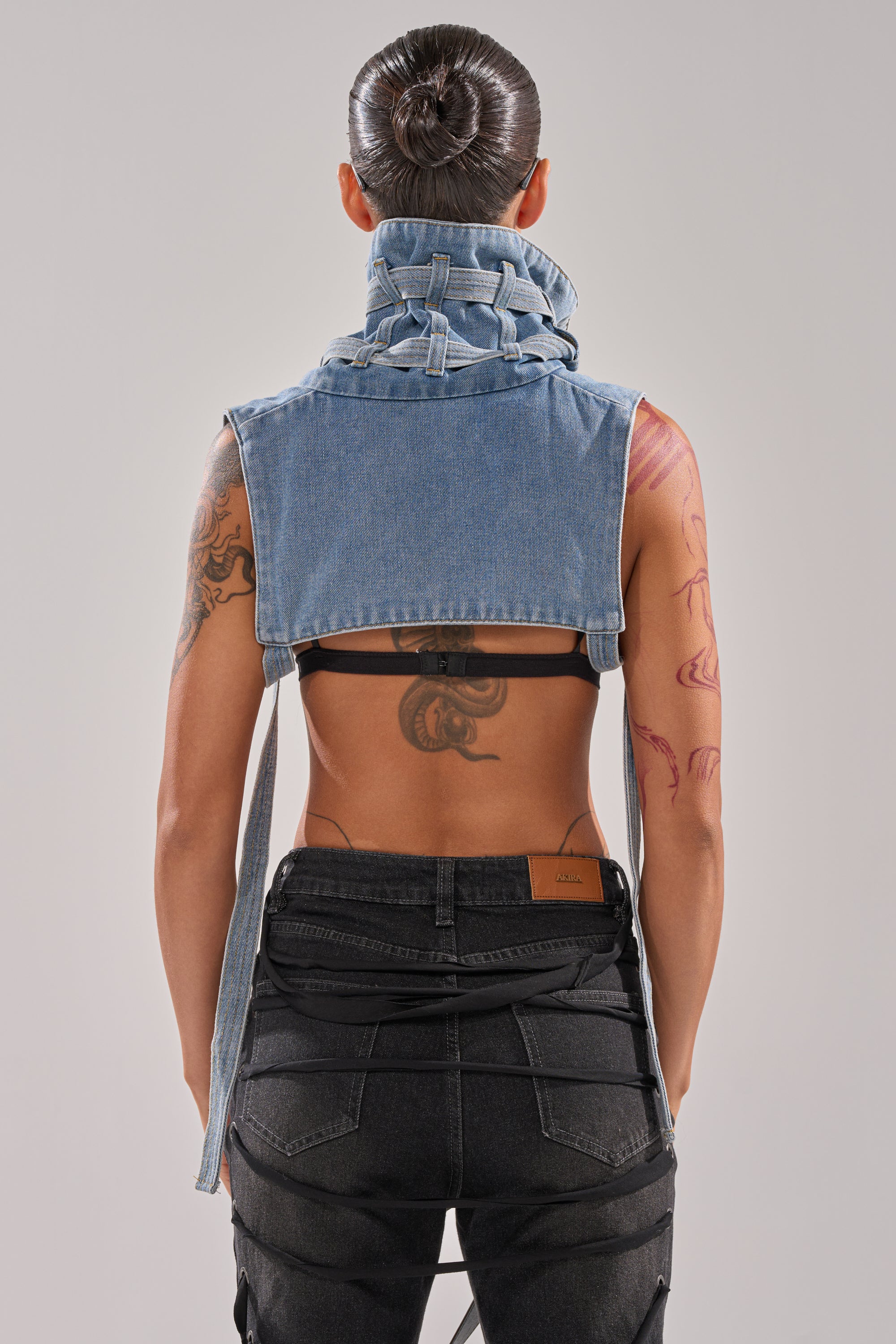 A person with tattoos and a sleek bun is seen from behind wearing the NO SHOW JACKET COLLAR, an ultra-cropped sleeveless washed denim vest, paired with black pants against a plain background.