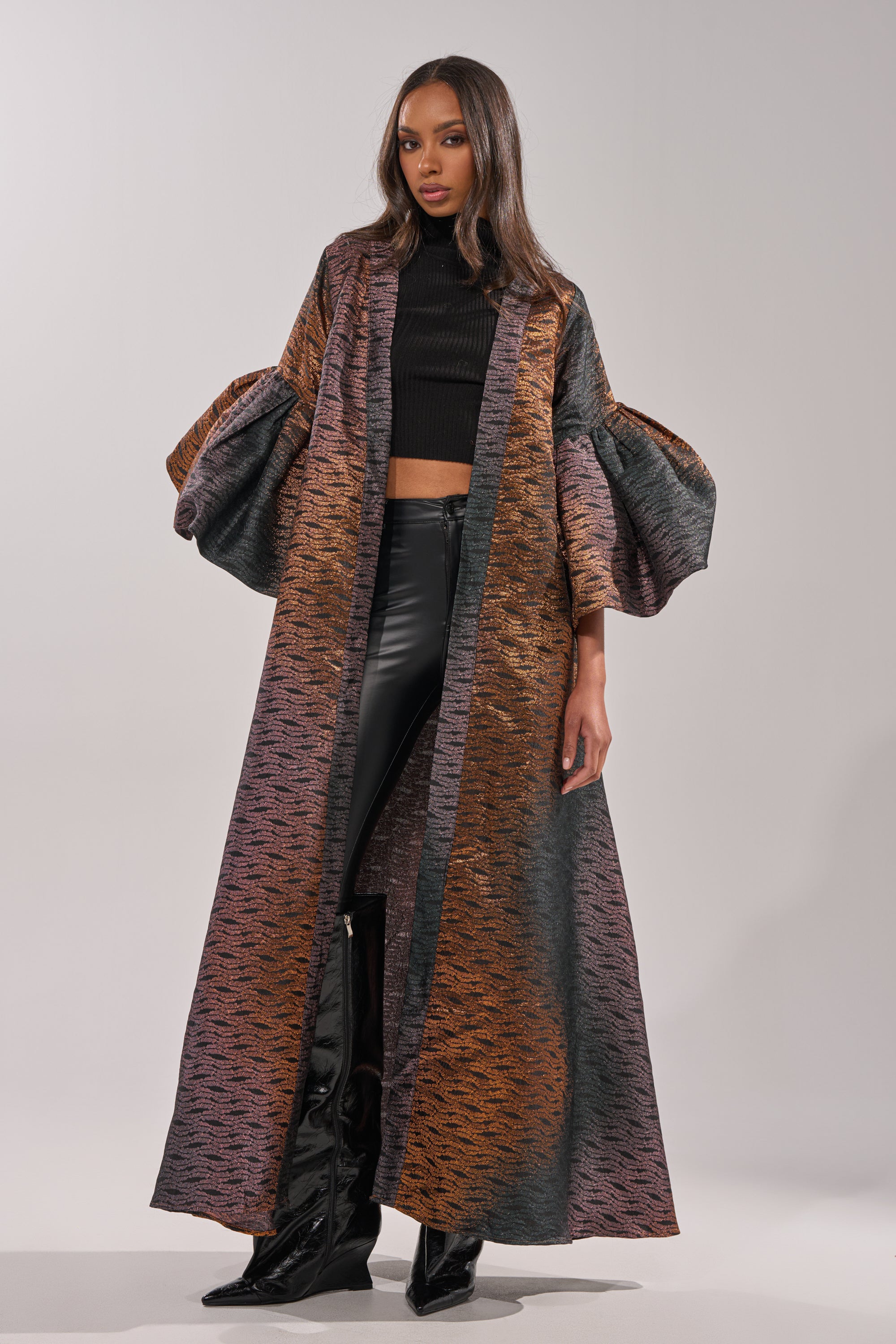 A woman stands against a plain light background wearing the FLORENCE BROCADE PUFF SLEEVE DUSTER over a black crop top and shiny black pants tucked into pointed boots. 0