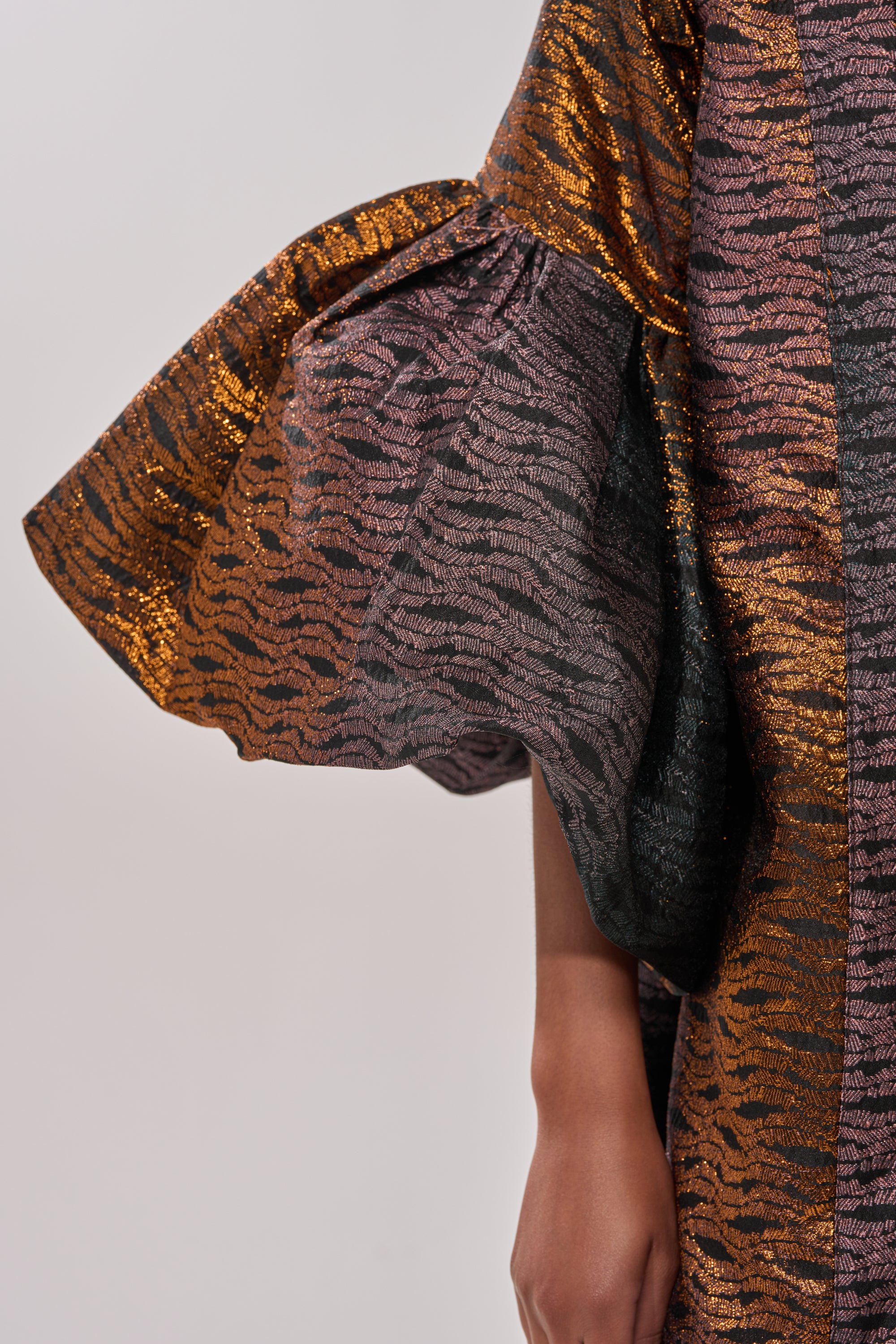 A close-up shows an arm wearing the FLORENCE BROCADE PUFF SLEEVE DUSTER—statement outerwear with dramatic metallic ruffled sleeves in bronze, gold, and black. The background is plain and light-colored. 9