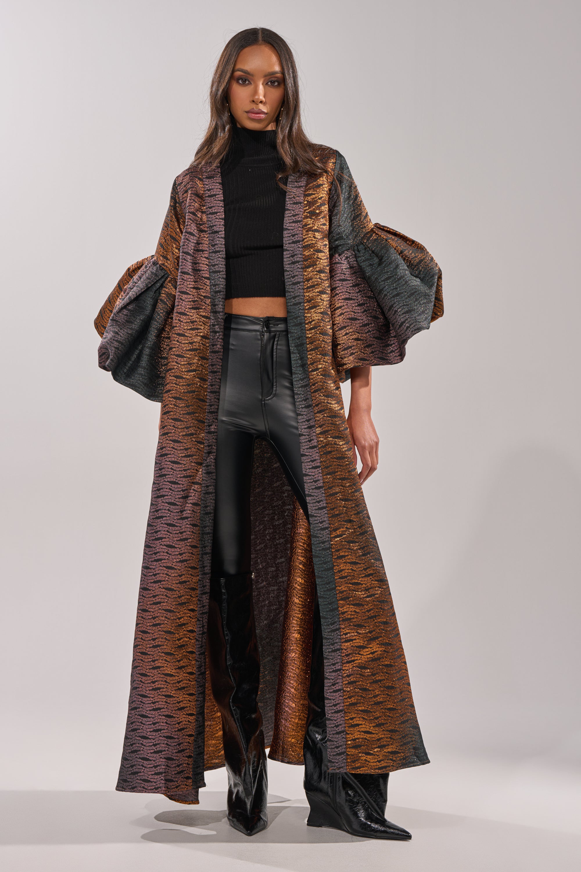 A woman stands confidently in the FLORENCE BROCADE PUFF SLEEVE DUSTER, layered over a black turtleneck crop top, black leather pants, and black heeled boots against a plain backdrop. 2