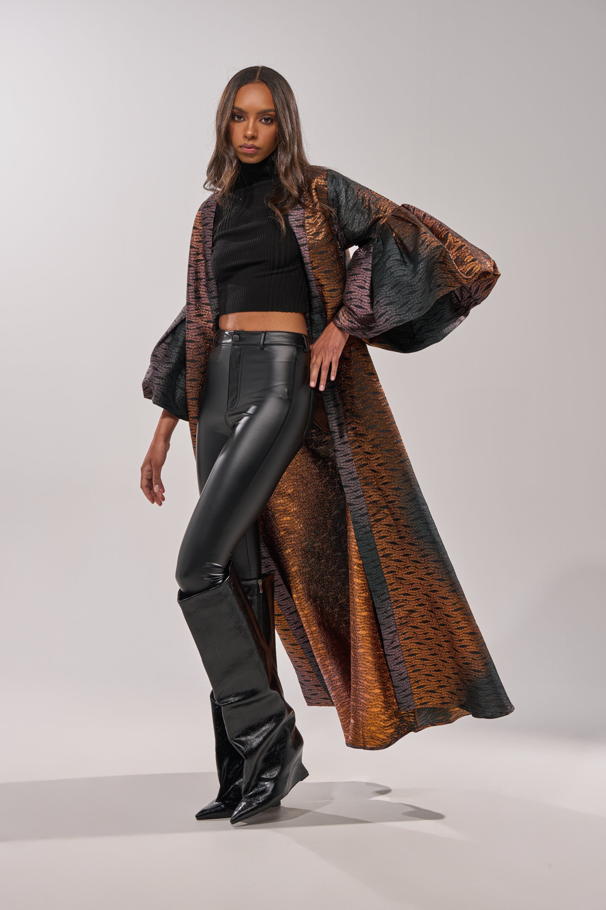 A woman poses confidently against a plain light gray background in black high-waisted leather pants, knee-high boots, a black crop top, and the FLORENCE BROCADE PUFF SLEEVE DUSTER—a long metallic outerwear piece with wide sleeves. 3