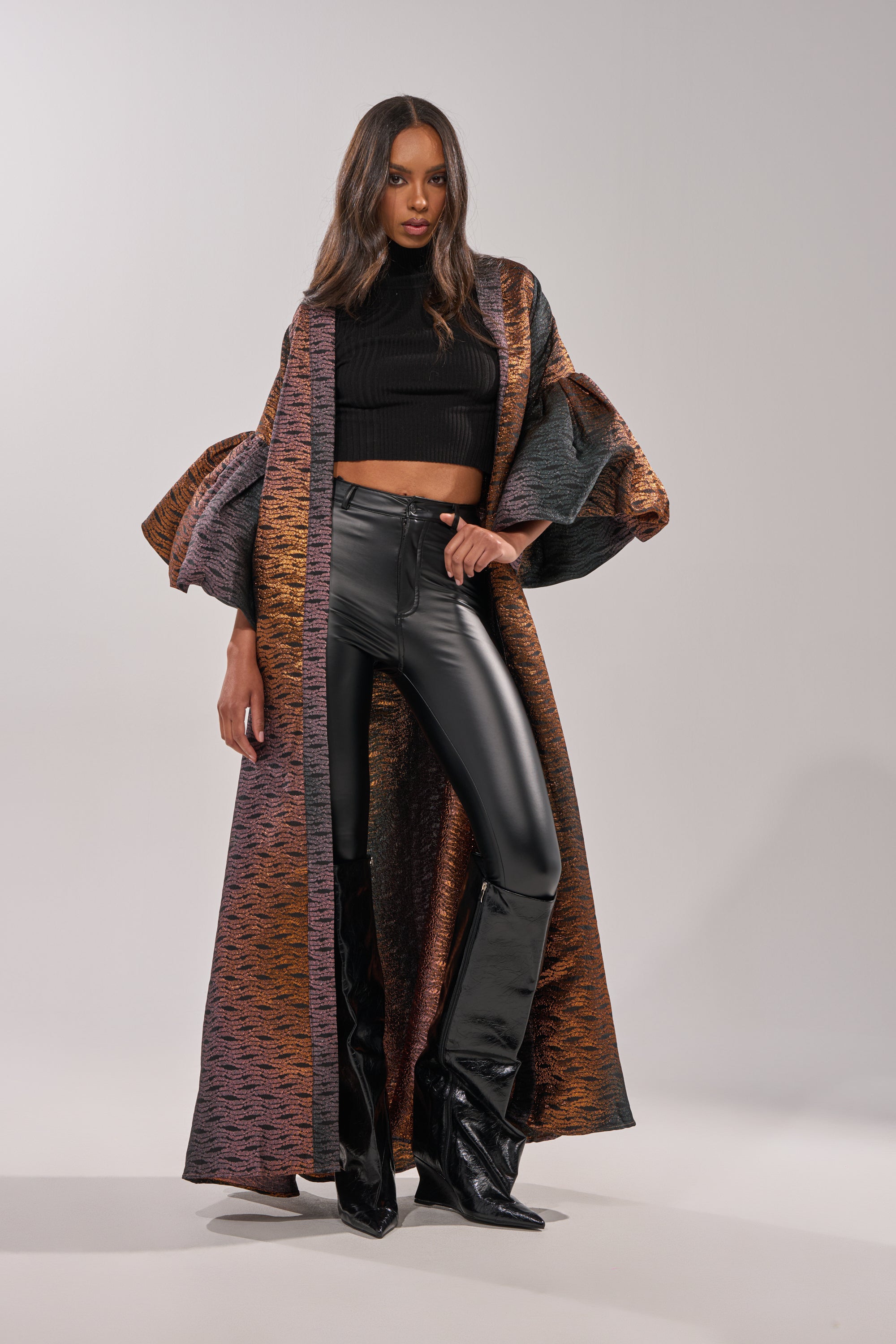 A woman poses confidently in the FLORENCE BROCADE PUFF SLEEVE DUSTER— a floor-length, multicolored metallic piece with dramatic puff sleeves—styled with a black crop top, high-waisted black leather pants, and heeled boots against a plain background. 4