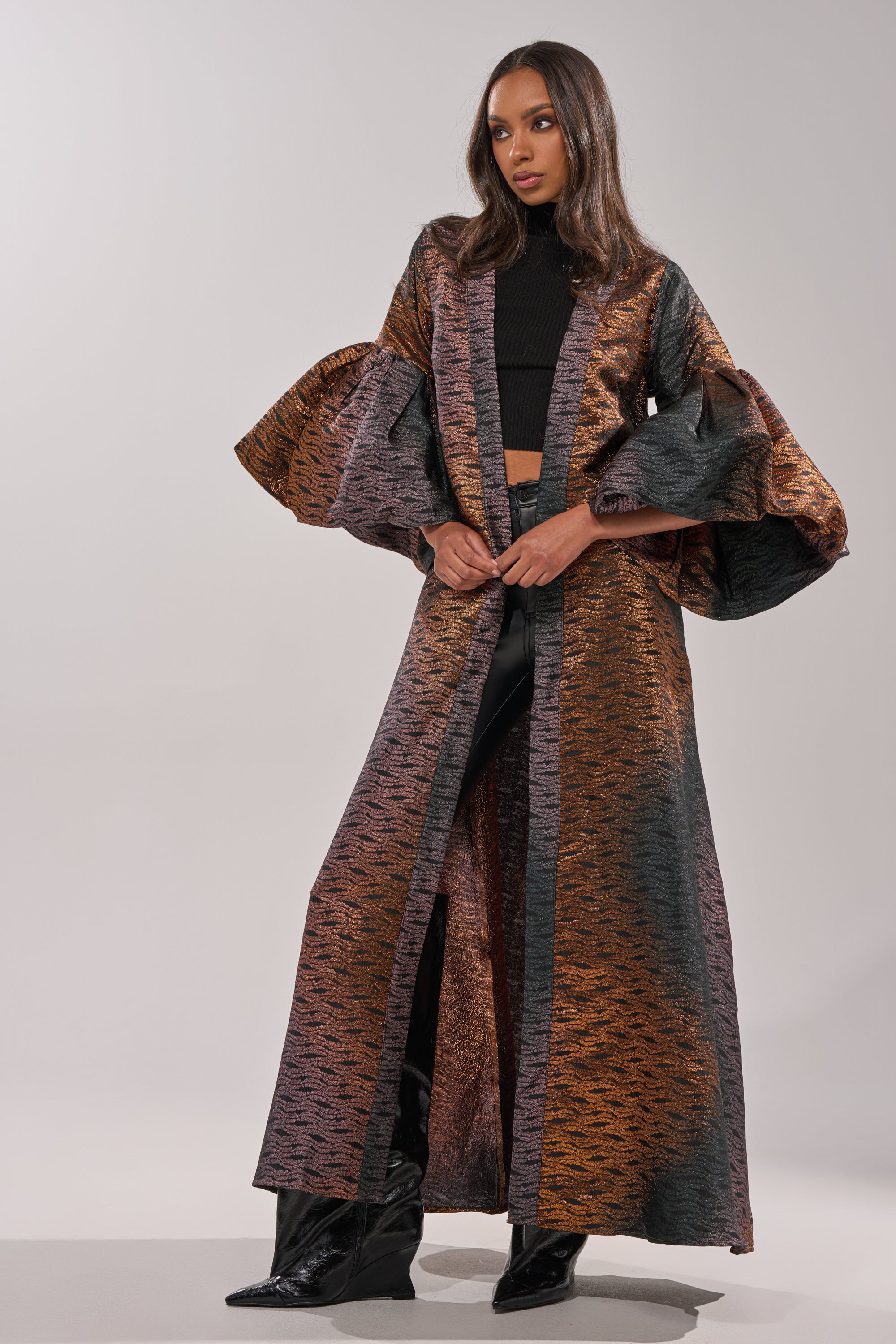 A woman wears the FLORENCE BROCADE PUFF SLEEVE DUSTER with dramatic ruffled sleeves over a black crop top and pants, styled with black heeled boots against a plain, light background. 5