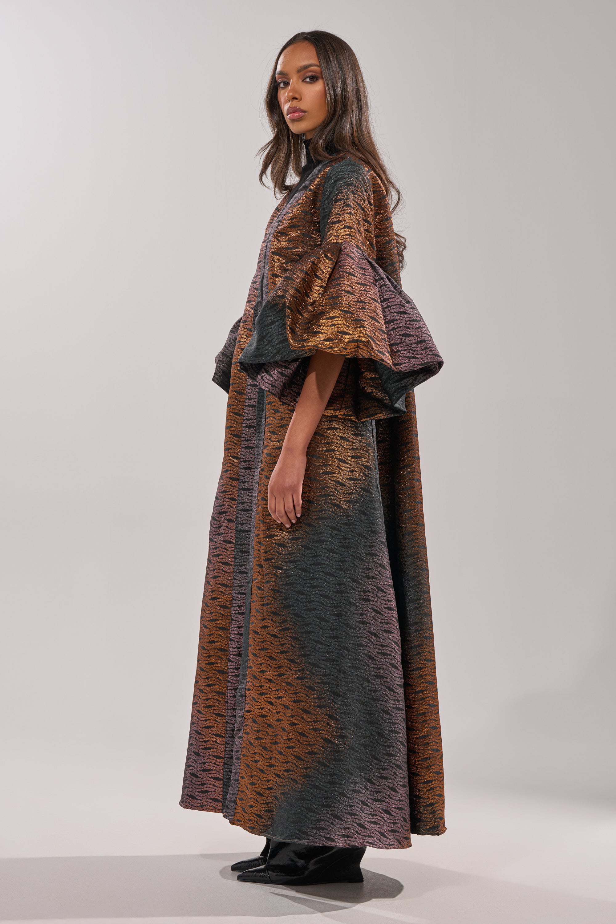 A woman with long, loose hair poses sideways against a plain light background wearing the FLORENCE BROCADE PUFF SLEEVE DUSTER, a floor-length dress in orange, purple, and dark gray with wide puff sleeves. 6