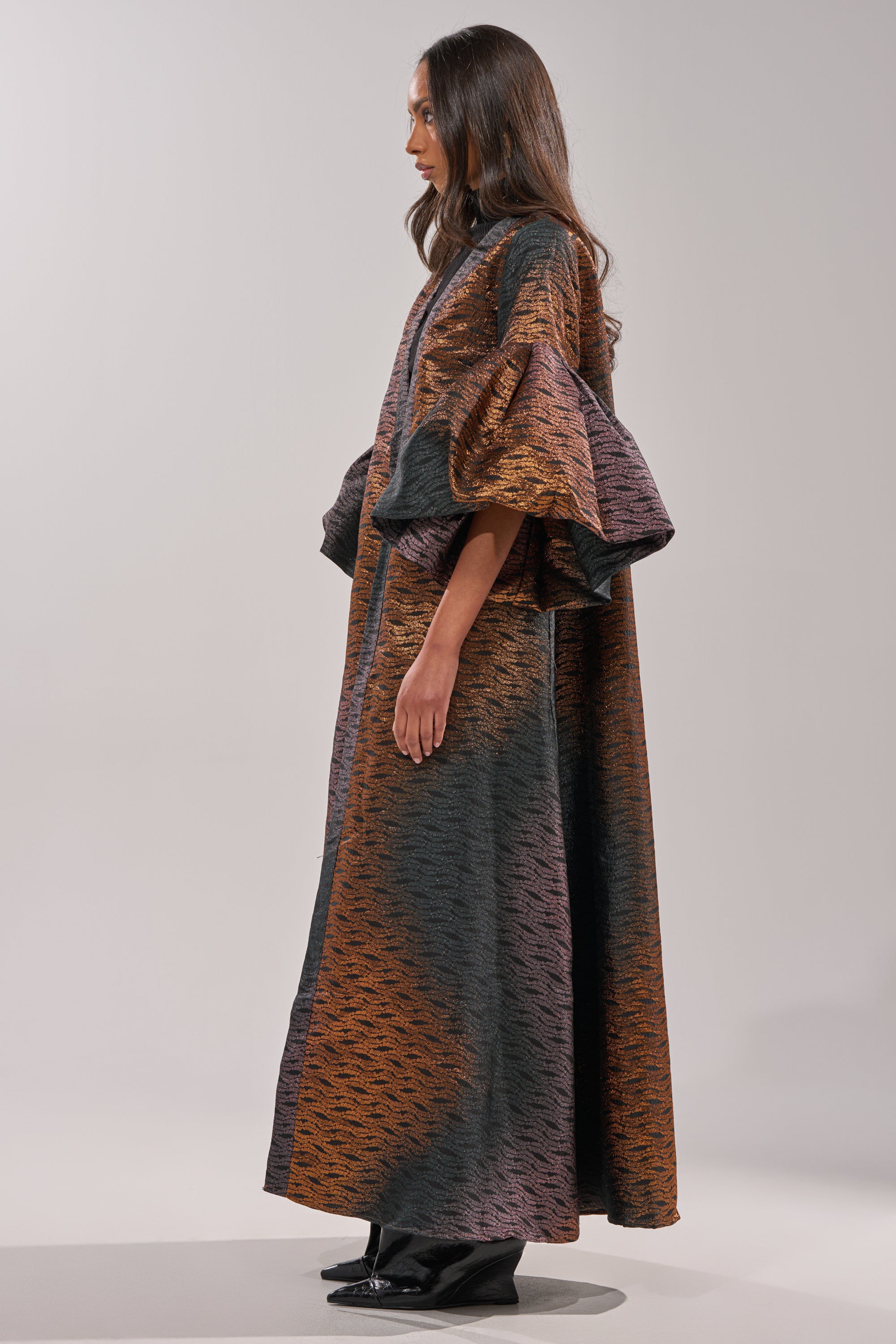 A person stands in profile, fashionably styled in the FLORENCE BROCADE PUFF SLEEVE DUSTER—a loose, floor-length piece with voluminous puff sleeves and rich dark orange, brown, and black metallic brocade tones. 7