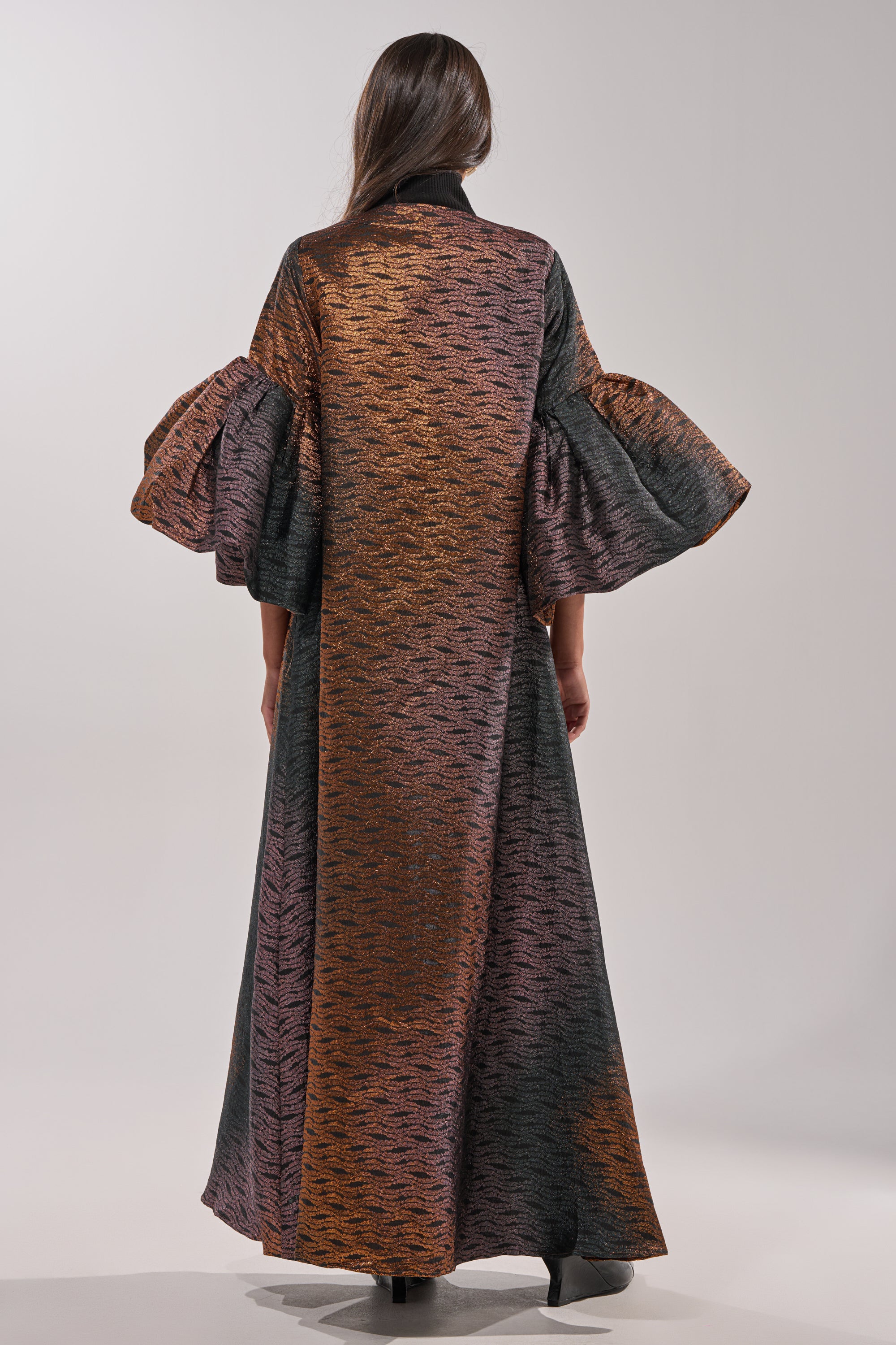 A woman faces away, her straight hair down her back, wearing the FLORENCE BROCADE PUFF SLEEVE DUSTER in dark bronze and copper metallic tones with wide, dramatic sleeves. She poses against a plain, light background. 8