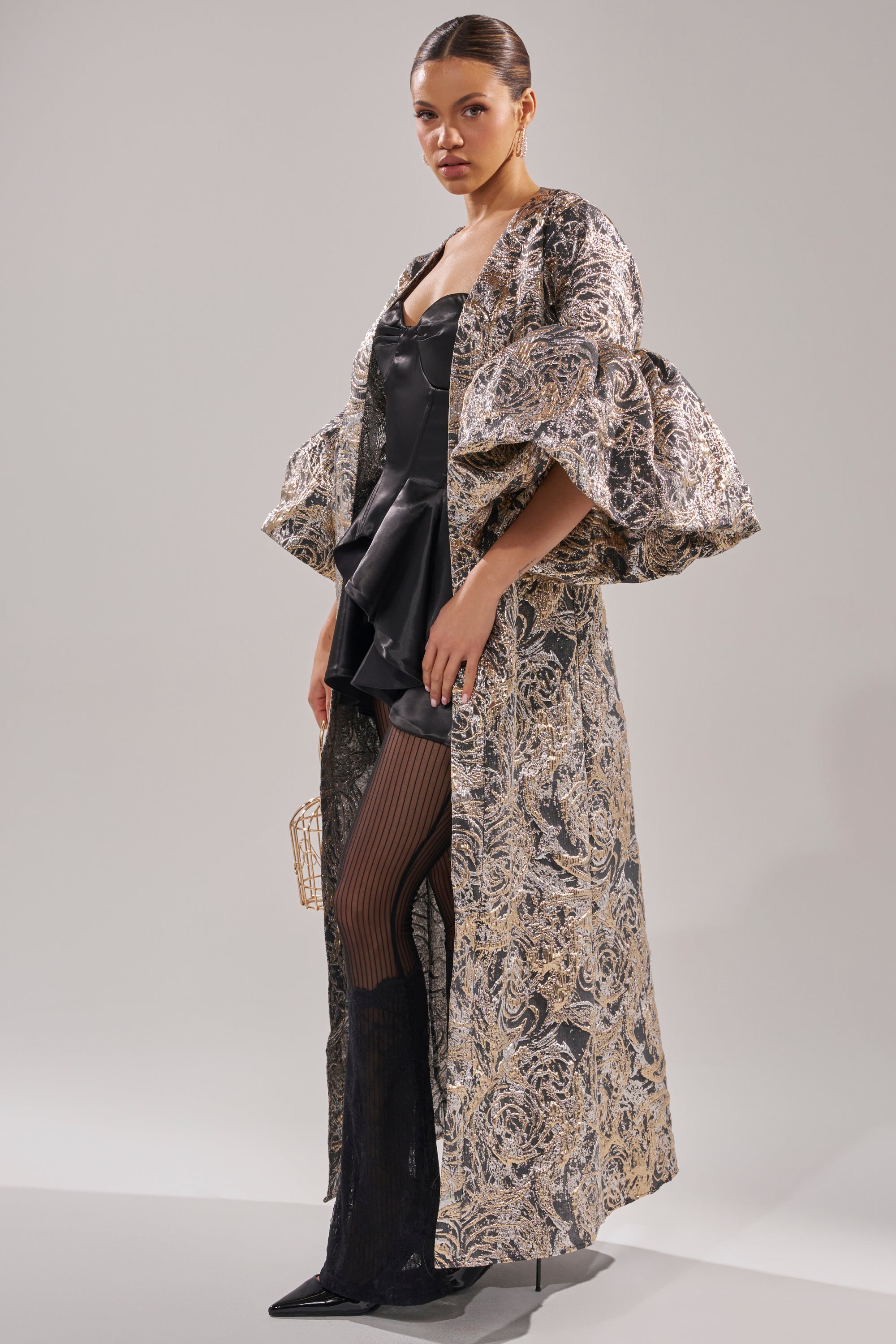 A woman poses in the METALLIC PUFF SLEEVE BROCADE DUSTER IN SILVER over a black corset dress, sheer striped tights, and pointed black boots, accessorizing with a small gold handbag. 0