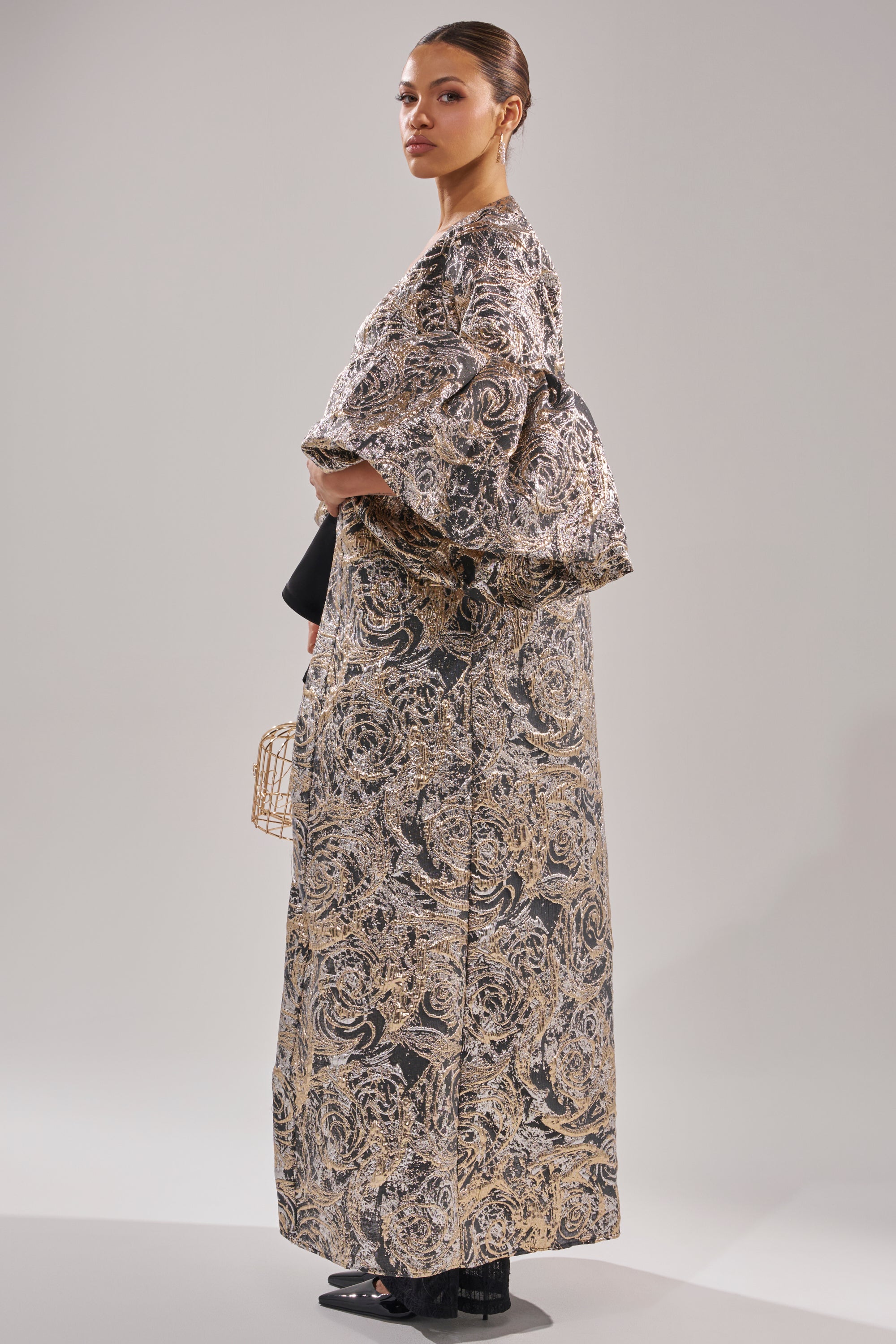 A person stands in profile wearing the METALLIC PUFF SLEEVE BROCADE DUSTER IN SILVER with wide sleeves, holding a geometric gold purse. They wear black shoes, have hair pulled back with large earrings, against a plain, light background. 2