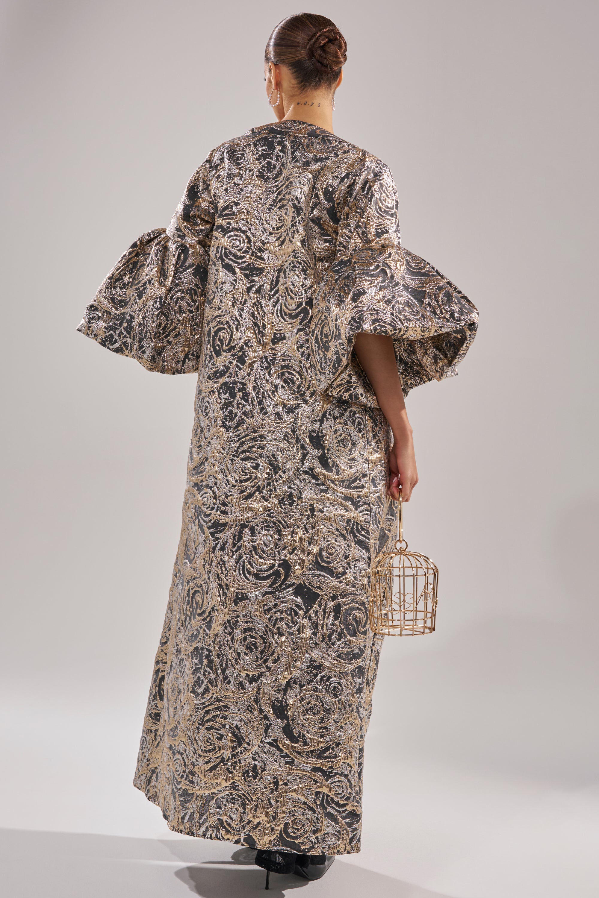 A person faces away, wearing the METALLIC PUFF SLEEVE BROCADE DUSTER IN SILVER with wide sleeves—a stunning holiday look. Their hair is in a neat bun, and they hold a small, gold cage-like purse in their left hand. 3
