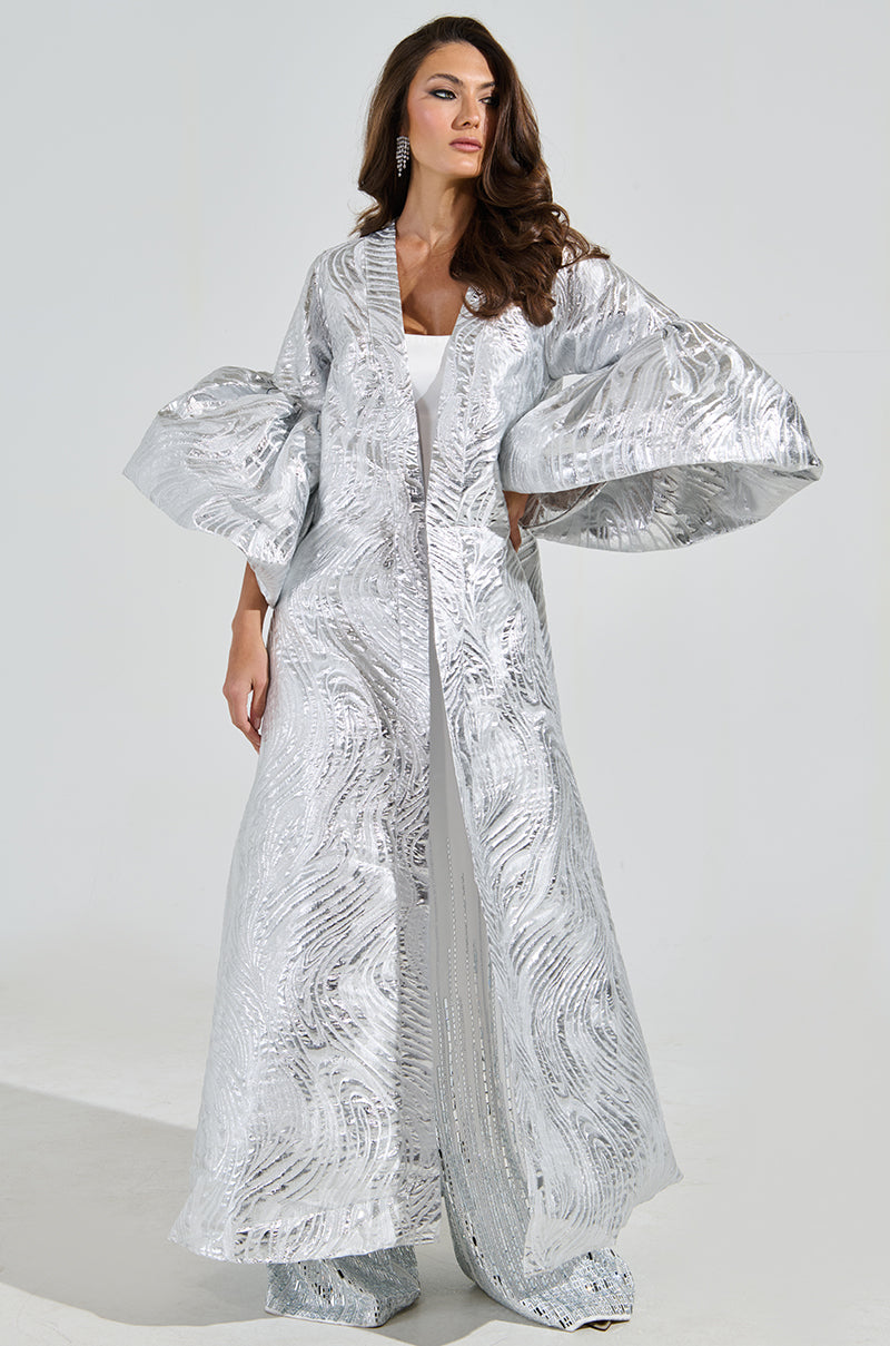A woman wears the Maldives Brocade Puff Sleeve Duster, a silver brocade-patterned floor-length robe with dramatic sleeves over a white outfit. Her long brown hair cascades down as she accessorizes with earrings, standing against a plain backdrop.