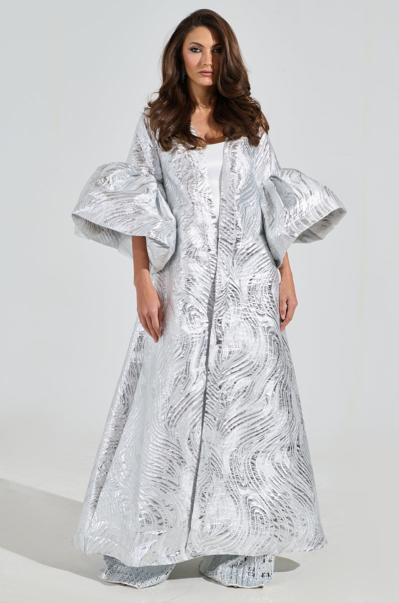 A woman wears the Maldives Brocade Puff Sleeve Duster, a shimmering silver gown with voluminous sleeves and swirling patterns. Her long, wavy brown hair frames her neutral expression against a plain backdrop.