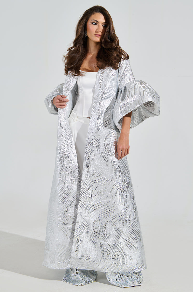 A woman with long brown hair stands confidently, wearing a shiny MALDIVES BROCADE PUFF SLEEVE DUSTER over a white outfit. The plain, light gray background enhances the metallic sheen of the coat's brocade pattern and puff sleeves.