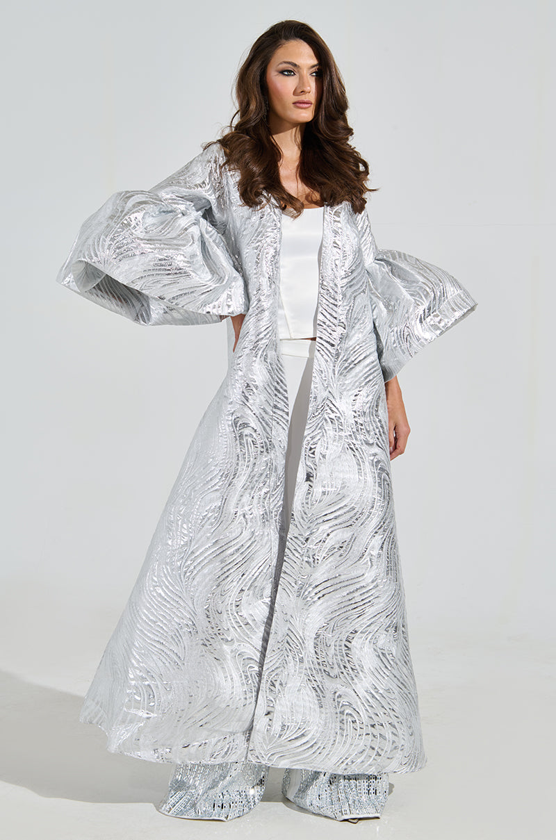 A woman poses against a plain background, wearing the MALDIVES BROCADE PUFF SLEEVE DUSTER in silver with intricate brocade patterns. Over a white outfit, her long, wavy brown hair complements the dramatic puff sleeves of the flowing gown.