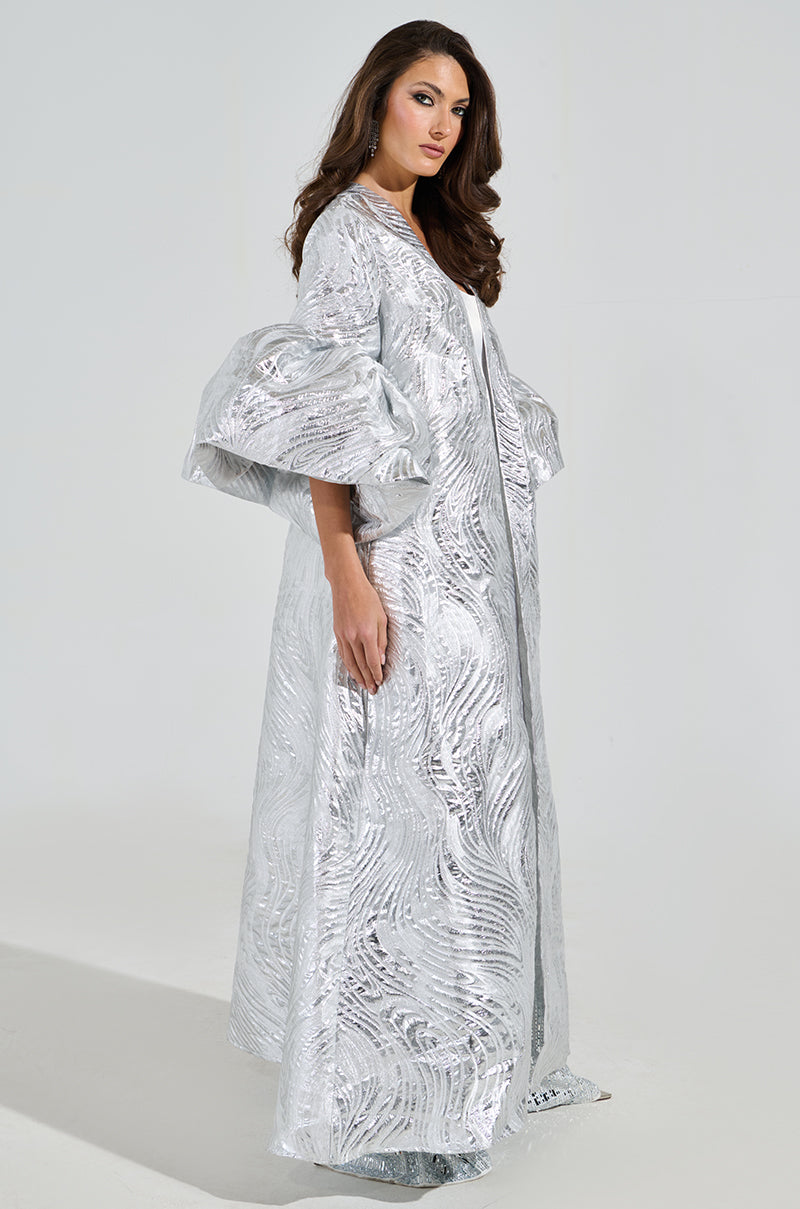 A woman poses against a plain background in the MALDIVES BROCADE PUFF SLEEVE DUSTER, showcasing its silver brocade pattern, voluminous structured sleeves, and flowing design. Her long wavy hair adds to her confident side profile stance.
