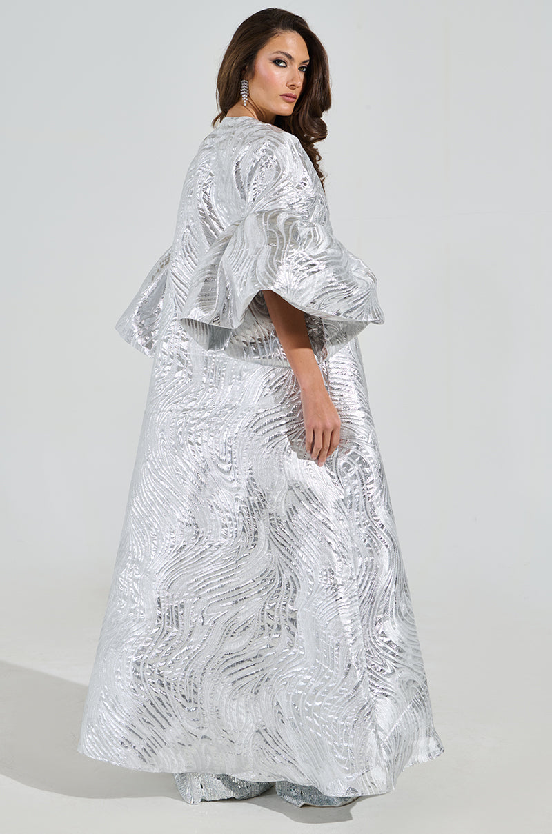 A woman confidently wears the elegant MALDIVES BROCADE PUFF SLEEVE DUSTER, a long silver gown with intricate metallic brocade patterns and voluminous sleeves. She poses gracefully against a plain background, one hand gently touching her hair.