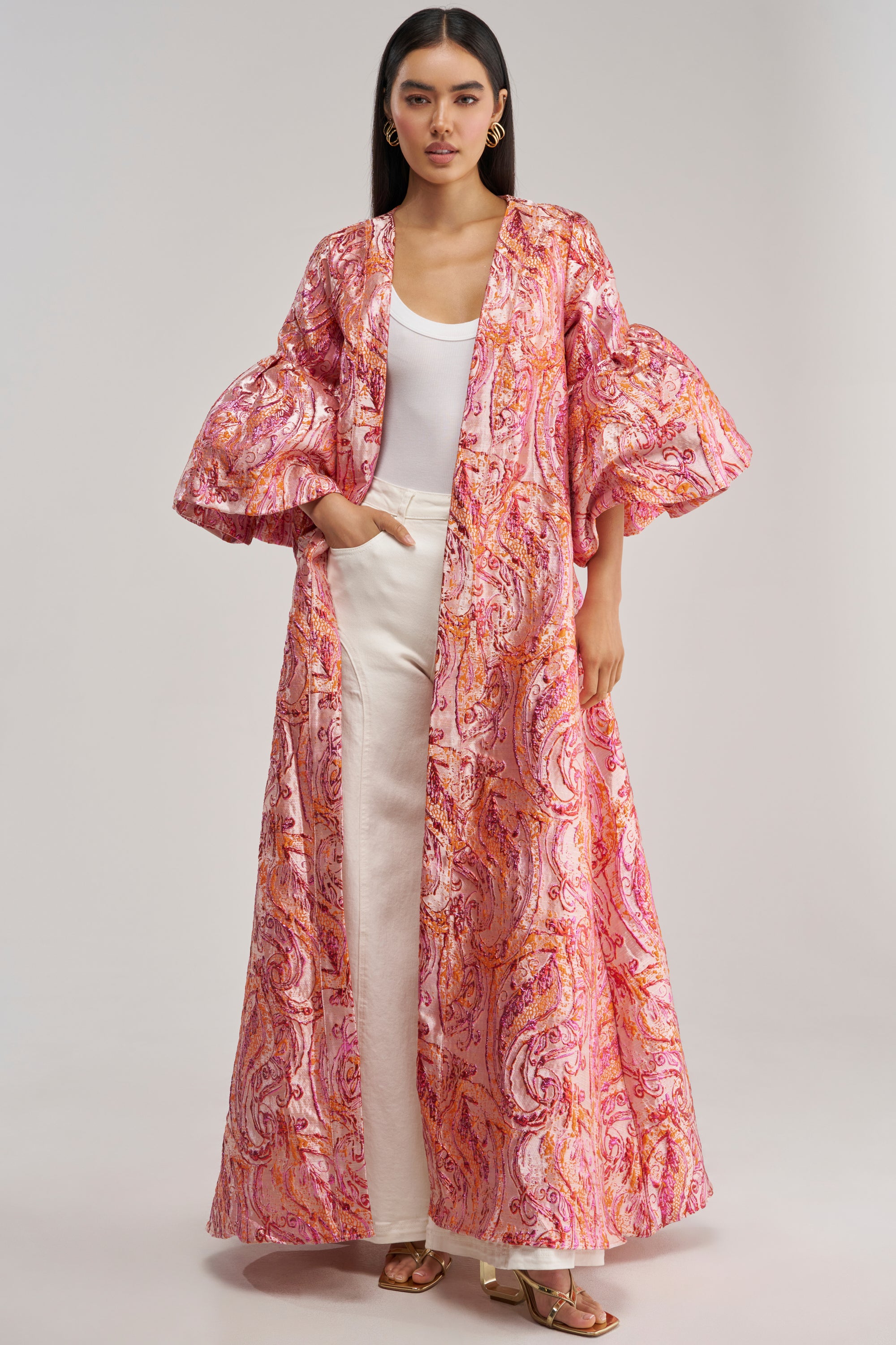A woman stands against a plain background, wearing the PINK SORBET BROCADE DUSTER—a vibrant hot pink and orange outerwear piece with a flowing, patterned design and puffy sleeves—layered over a white top and pants.