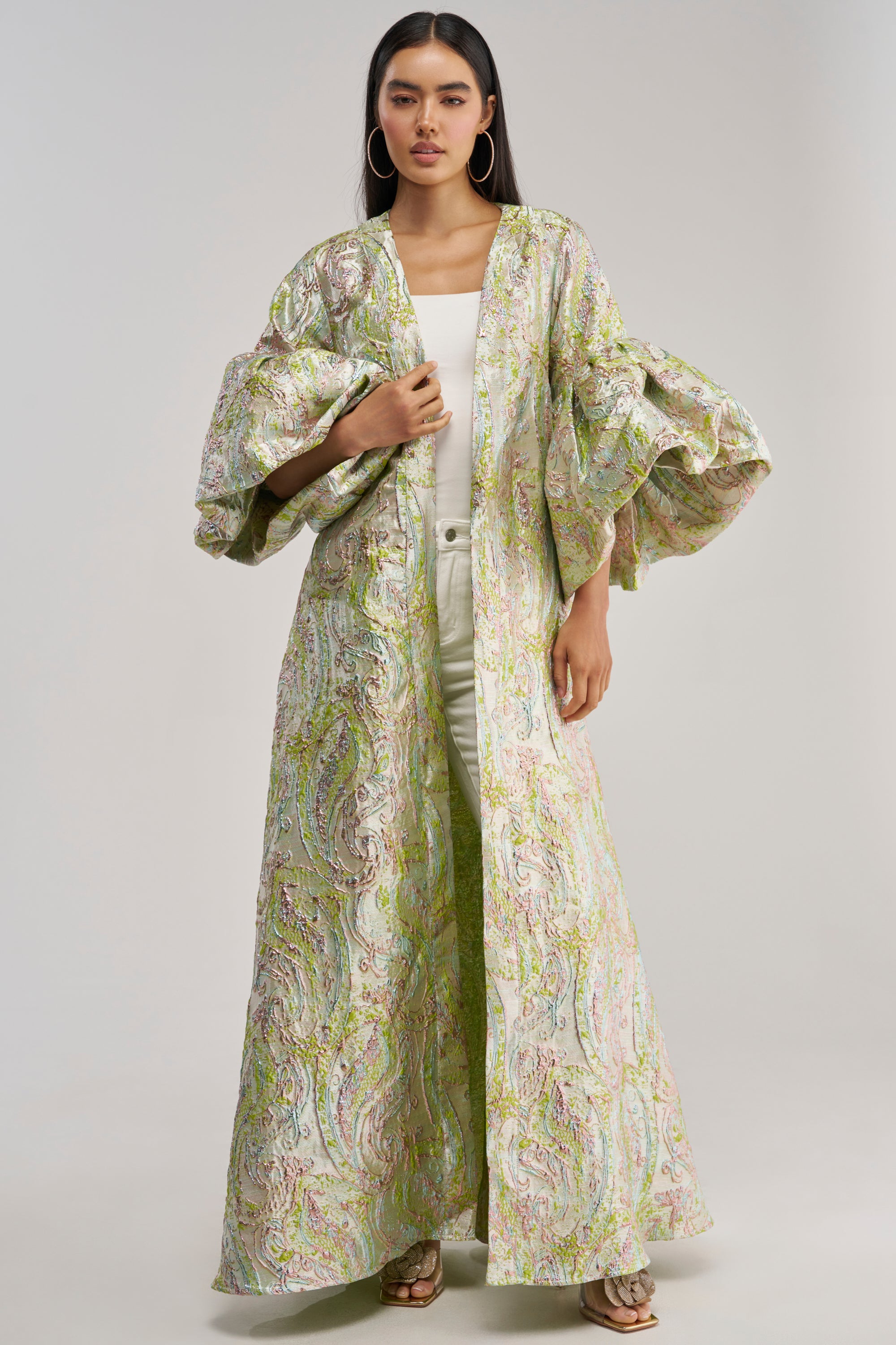 A person wears the KEY LIME BROCADE DUSTER—a floor-length, light-colored brocade piece with voluminous paisley-patterned sleeves—over a white top and pants, standing against a plain background.