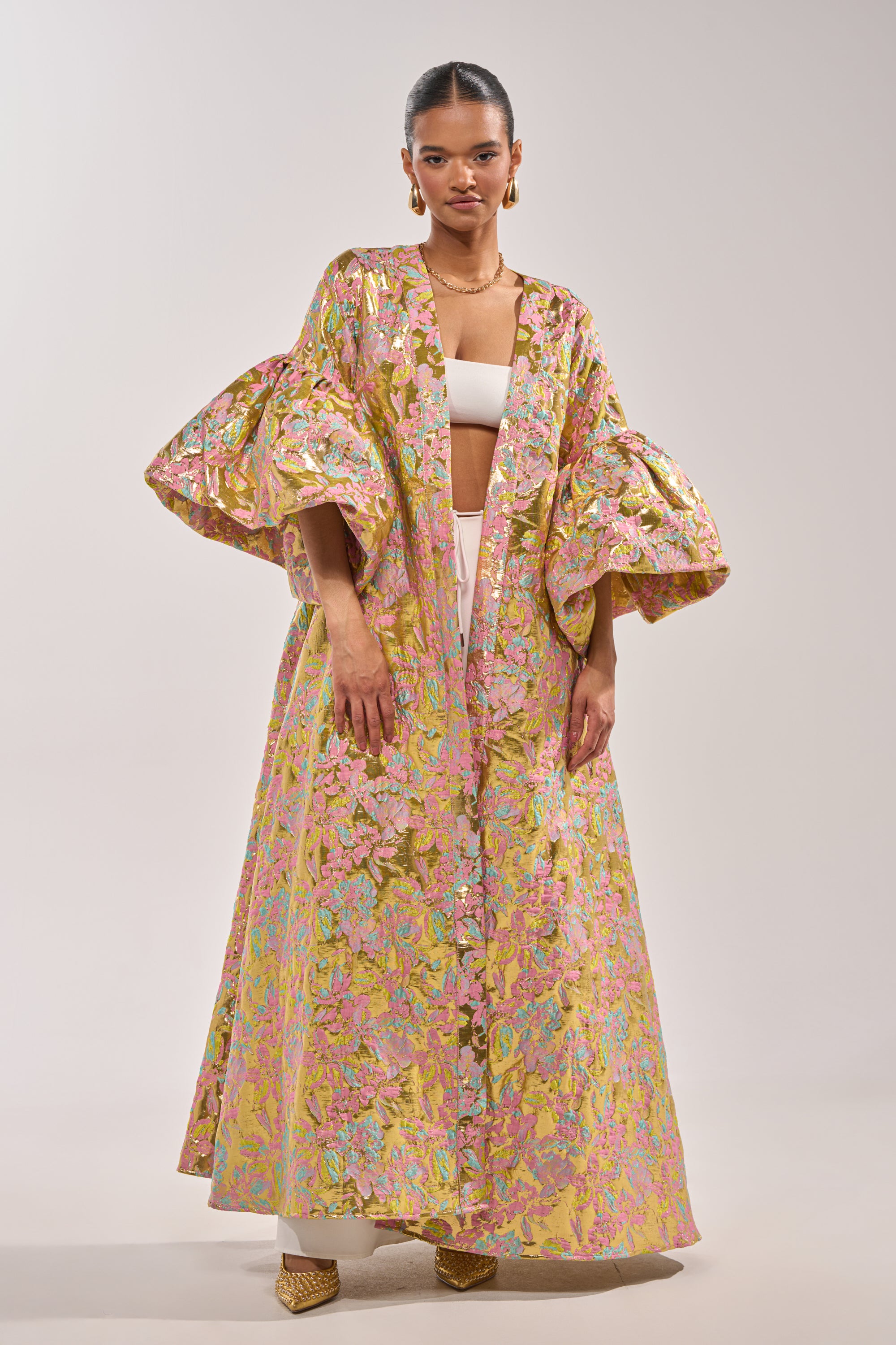 A woman models the LEMON DROP BROCADE DUSTER, featuring metallic pink, gold, and green hues with voluminous sleeves. She pairs it with a pastel top, white pants, gold earrings, and pointed shoes for a standout look. 0
