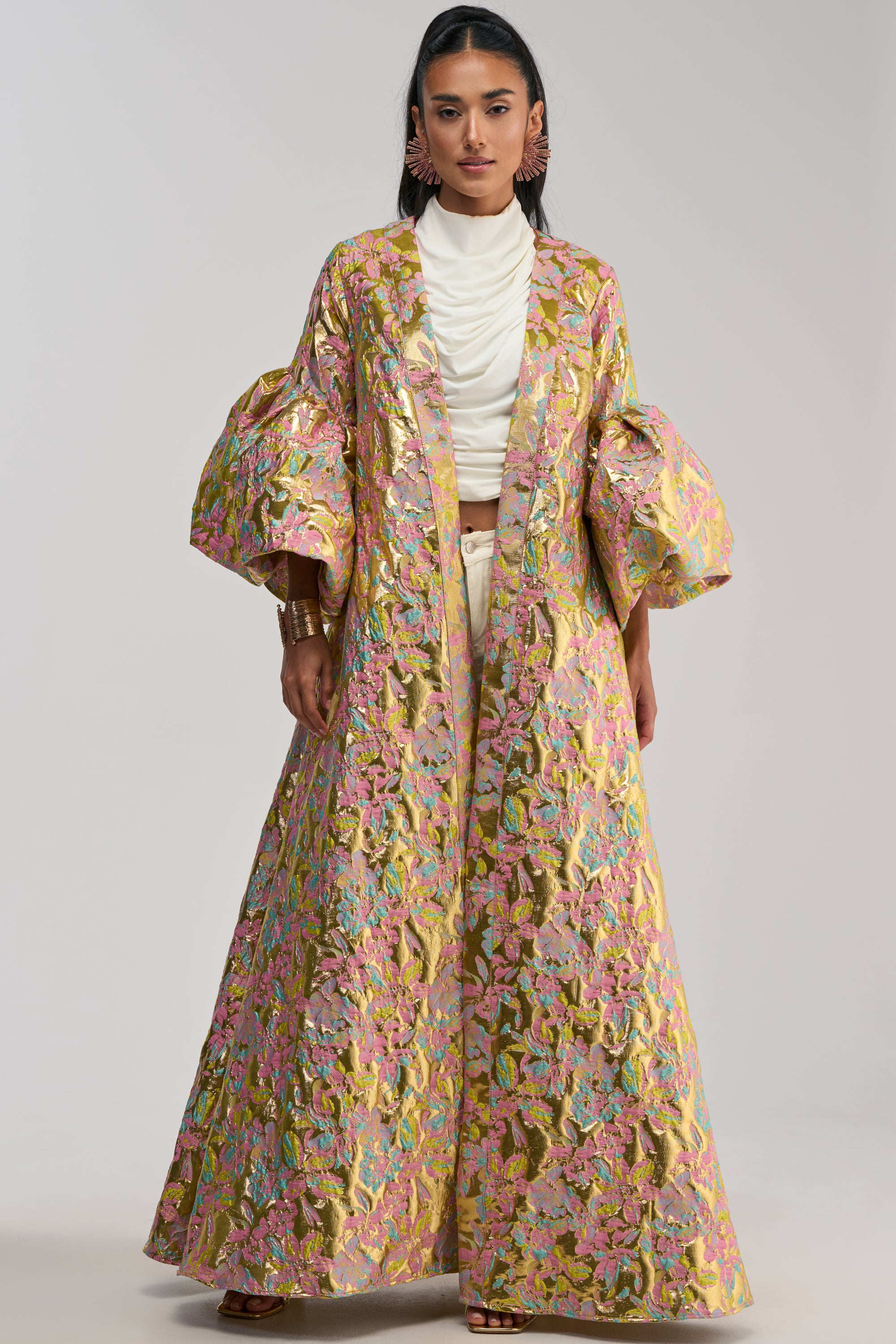 A woman wears the LEMON DROP BROCADE DUSTER—a long, colorful floral brocade coat with wide, puffed sleeves—over a white ruched statement top and cream pants, standing against a plain light background.
