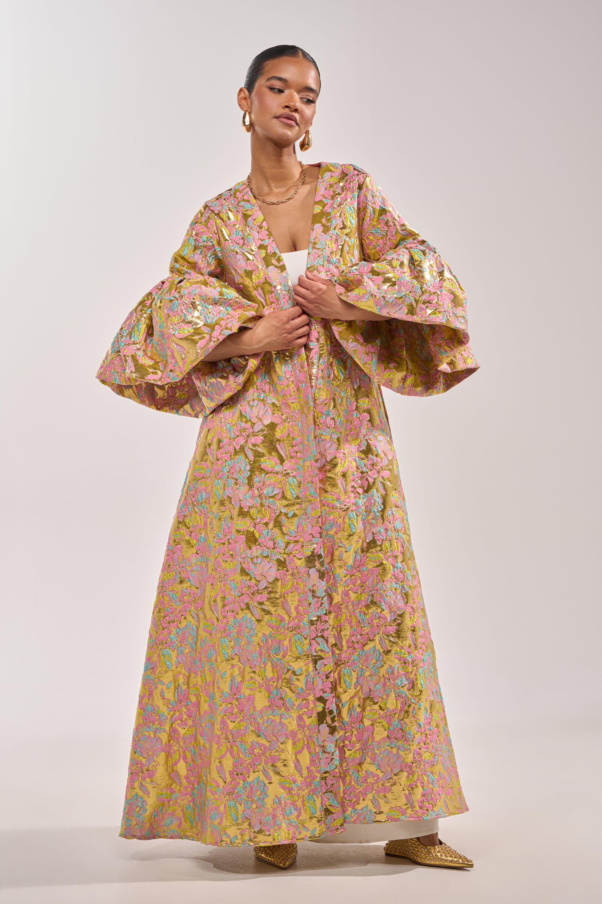 A woman models the LEMON DROP BROCADE DUSTER, a long, metallic piece with voluminous ruffled sleeves. Its pink, gold, and green patterns shine as she pairs it with gold hoop earrings, a necklace, and pointed shoes against a plain background. 1