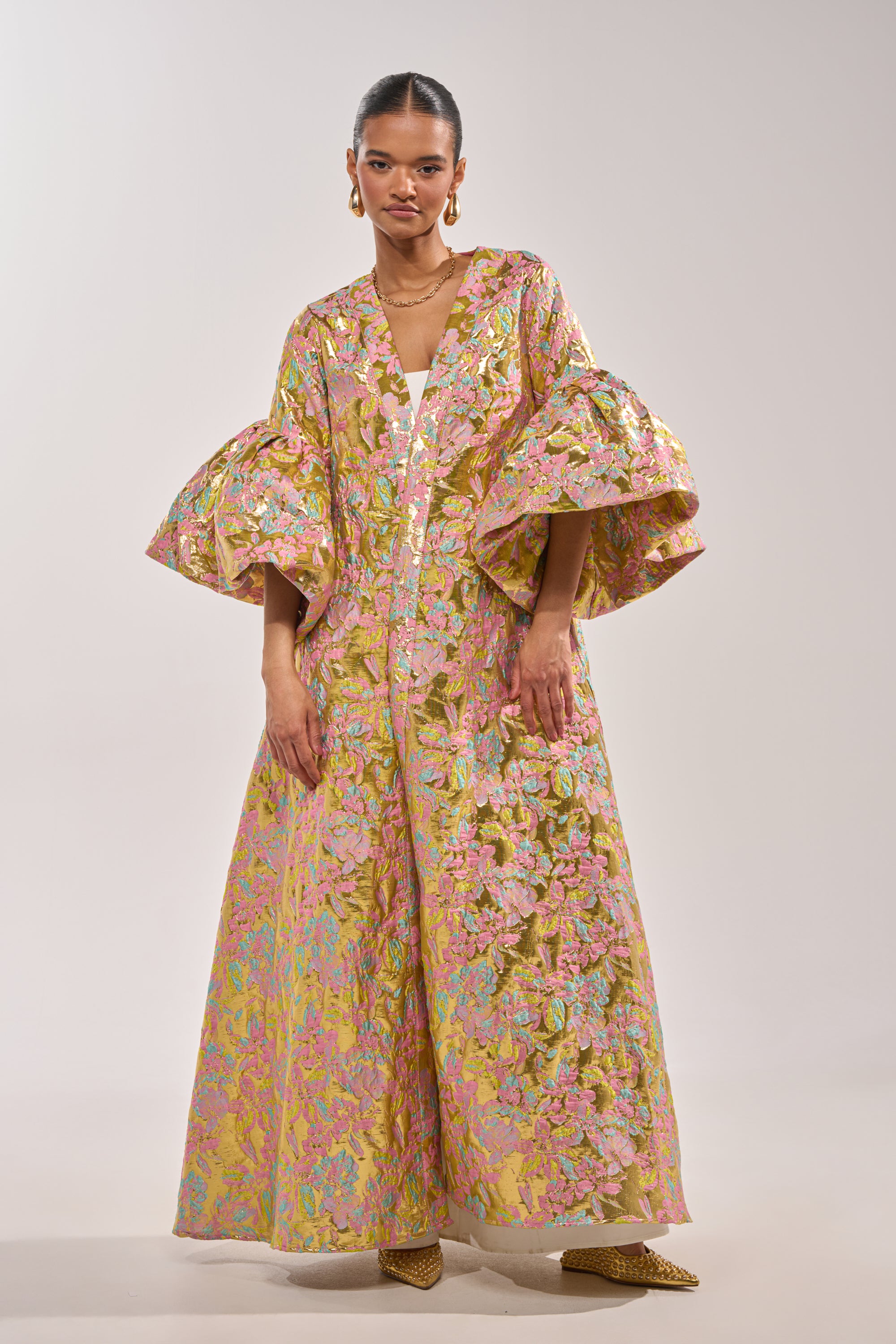 A woman wears the LEMON DROP BROCADE DUSTER, a long metallic floral brocade dress in pink, gold, and green with voluminous sleeves. She completes her look with slicked-back hair, gold earrings, a necklace, and statement gold heels. 2