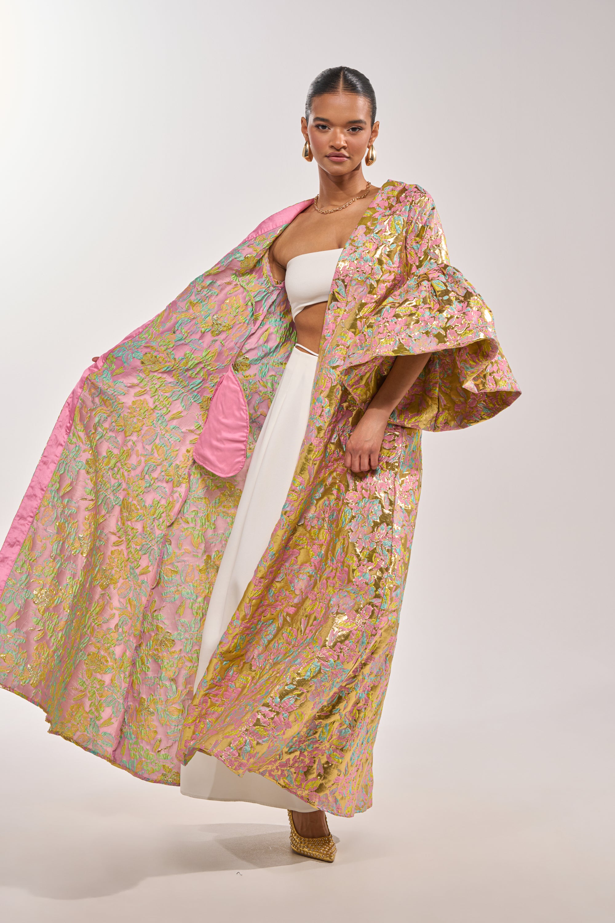 A woman poses in the LEMON DROP BROCADE DUSTER with pink lining over a white strapless top and wide-leg pants. Gold earrings and heels complement the statement layer, which stands out against a plain light background. 3