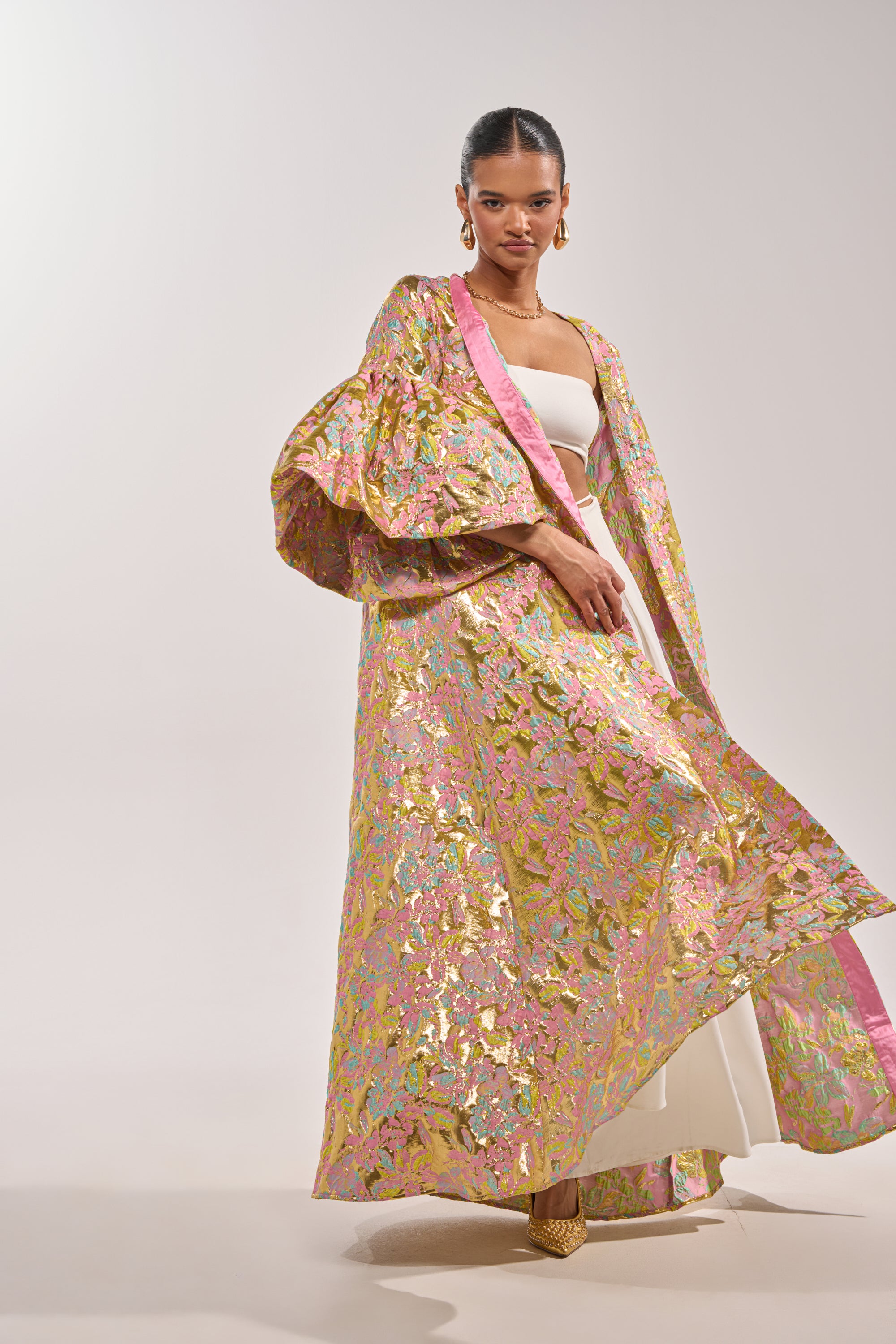 A woman stands confidently in the LEMON DROP BROCADE DUSTER with voluminous sleeves over a cream outfit, styled with hoop earrings, gold heels, and slicked-back hair against a plain light background. 4