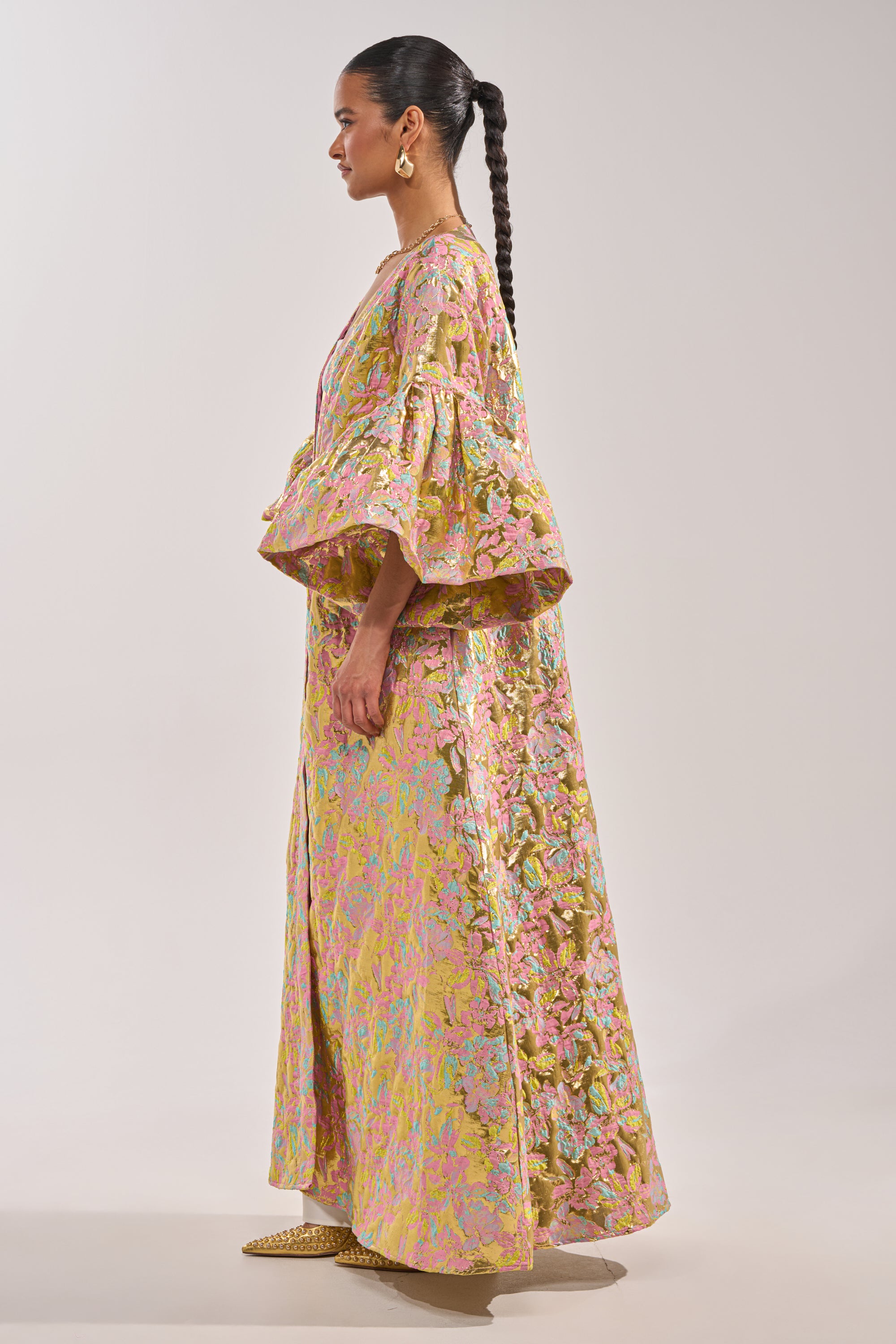 A woman in profile wears the LEMON DROP BROCADE DUSTER with flared sleeves and a long skirt. Her look is finished with a braided ponytail, gold earrings, and pointed shoes against a plain, light background. 5