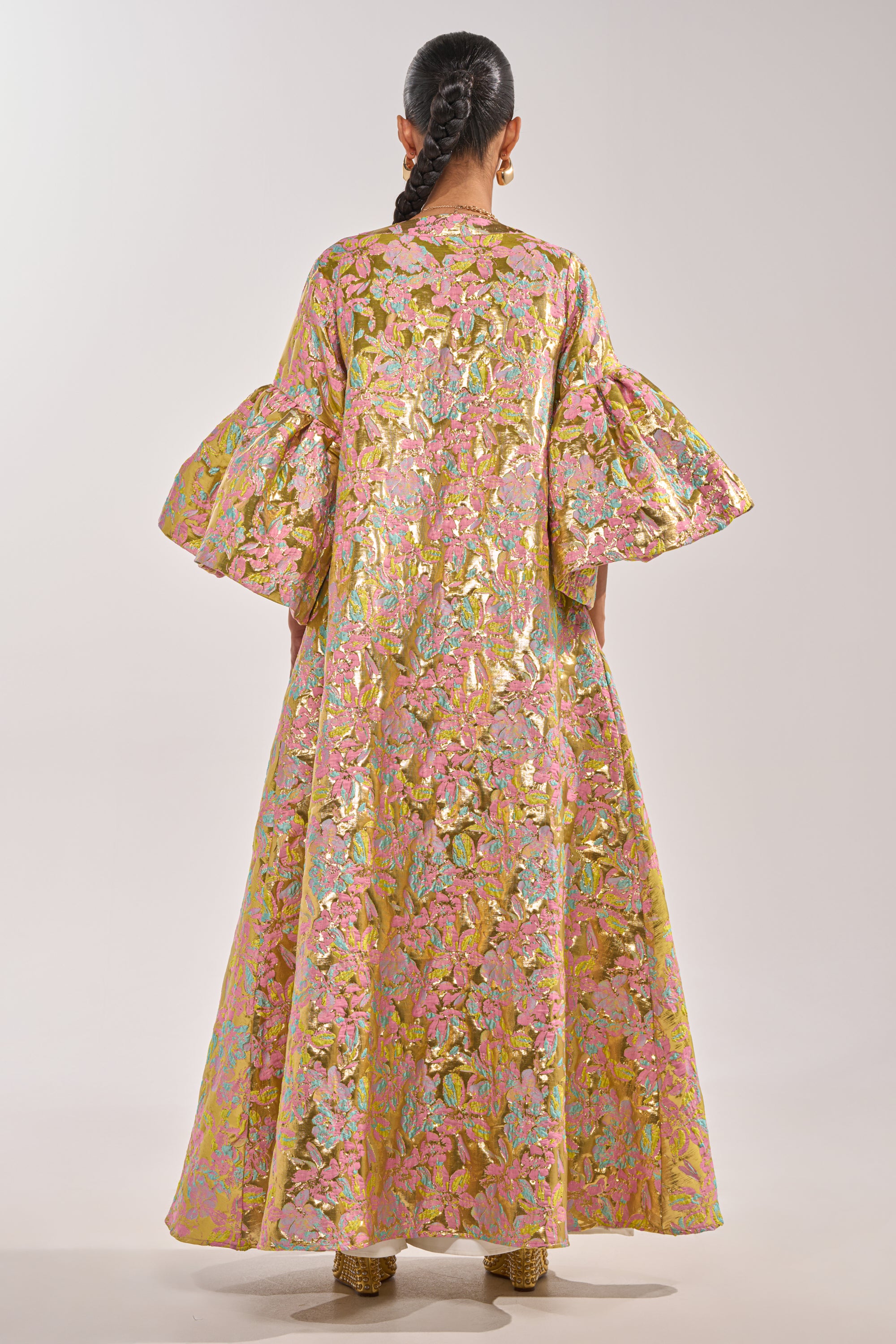 A person faces away wearing the LEMON DROP BROCADE DUSTER, a metallic floral layer in pink, gold, and green with voluminous sleeves. Styled with a long braid, gold earrings, and gold shoes for a bold statement look. 6