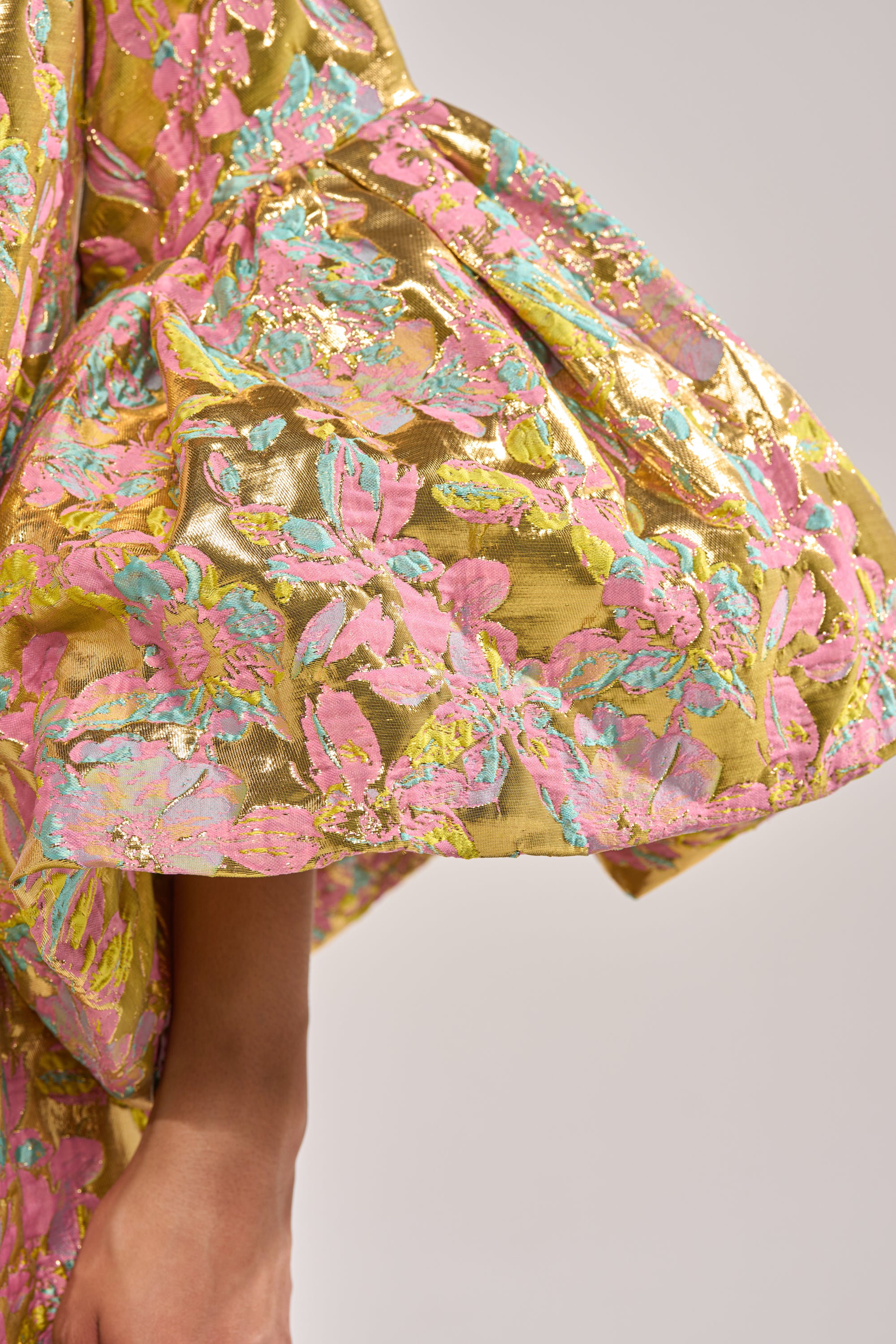 A close-up of the LEMON DROP BROCADE DUSTER shows its shiny gold metallic fabric with pink, blue, and green floral patterns. The textured material looks luxurious and bold, with the wearer's hand just slightly visible in the shot. 7