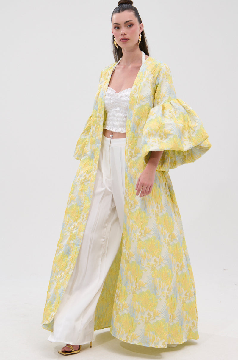 A woman stands confidently in the VENICE BROCADE PUFF SLEEVE DUSTER, a long yellow and white floral piece with voluminous sleeves, paired with white wide-legged pants and a cropped top against a plain white background.