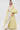 A woman stands confidently in the VENICE BROCADE PUFF SLEEVE DUSTER, a long yellow and white floral piece with voluminous sleeves, paired with white wide-legged pants and a cropped top against a plain white background.