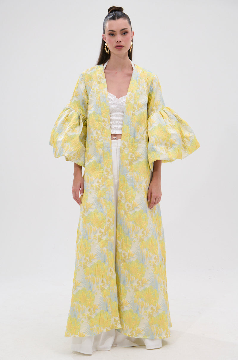 A woman wears the VENICE BROCADE PUFF SLEEVE DUSTER—long, yellow and blue floral brocade with voluminous sleeves—over a white lace top and white pants, standing against a plain white background.