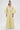 A woman wears the VENICE BROCADE PUFF SLEEVE DUSTER—long, yellow and blue floral brocade with voluminous sleeves—over a white lace top and white pants, standing against a plain white background.
