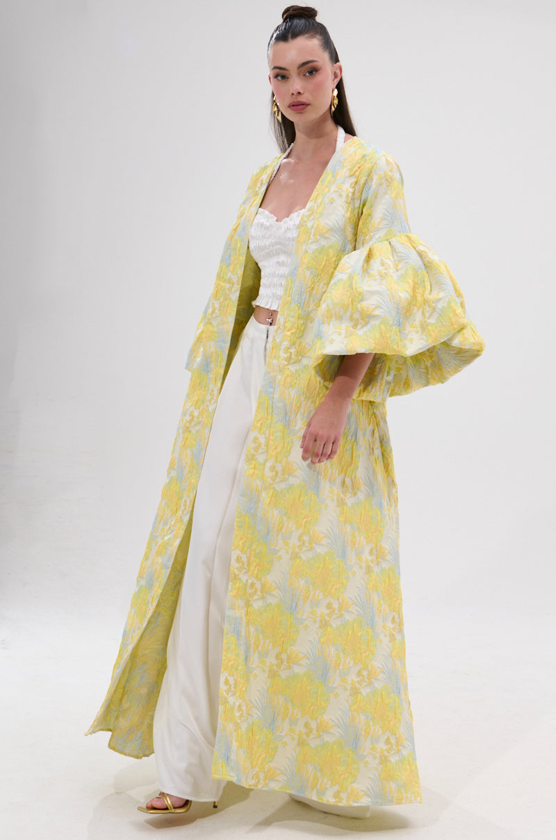 A woman wears the VENICE BROCADE PUFF SLEEVE DUSTER, a long yellow and light blue floral kimono with layered sleeves, over a white lace crop top and white pants, radiating vintage charm against a plain white background.