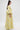 A woman in profile wears the VENICE BROCADE PUFF SLEEVE DUSTER—a long, yellow and white floral brocade dress with dramatic puff sleeves. She has long straight hair, hoop earrings, and poses against a plain white background.